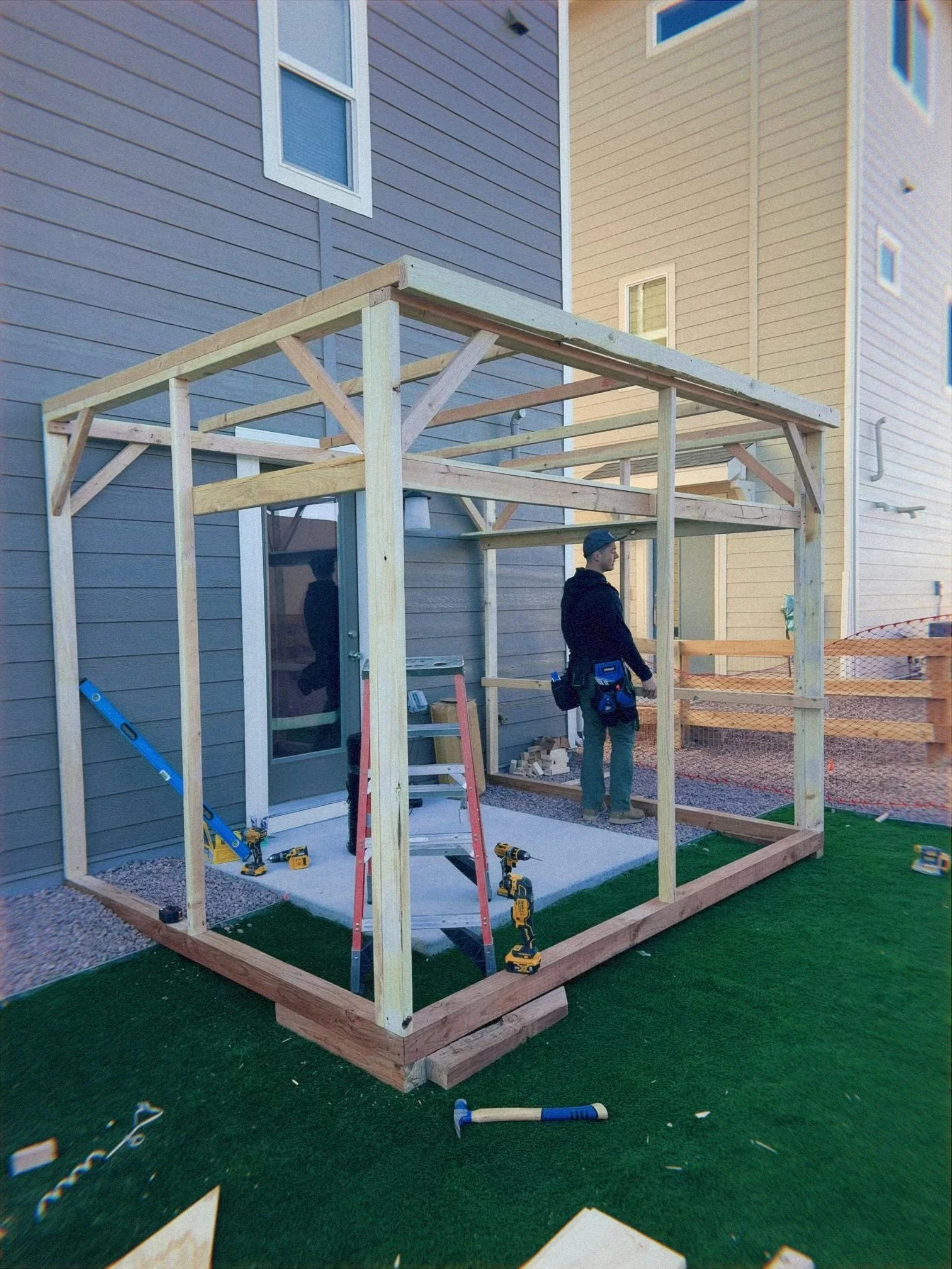 A man is working on constructing a wooden frame or deck attached to a house. The frame is in progress with some tools and materials on the ground nearby.