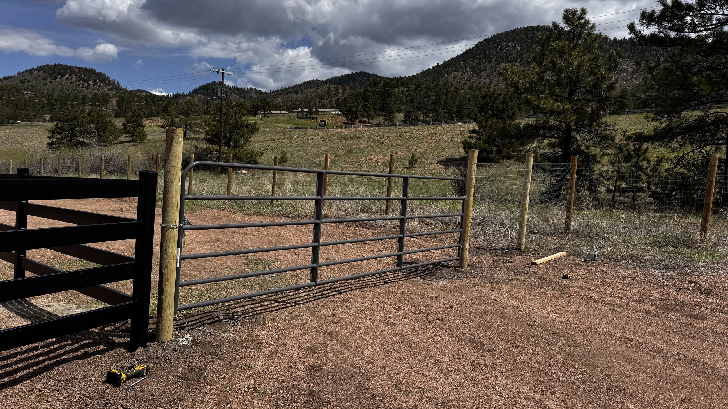 Construction site with metal gate and wooden posts, tools on reddish dirt ground, scenic background of green trees on hills, partly cloudy sky.