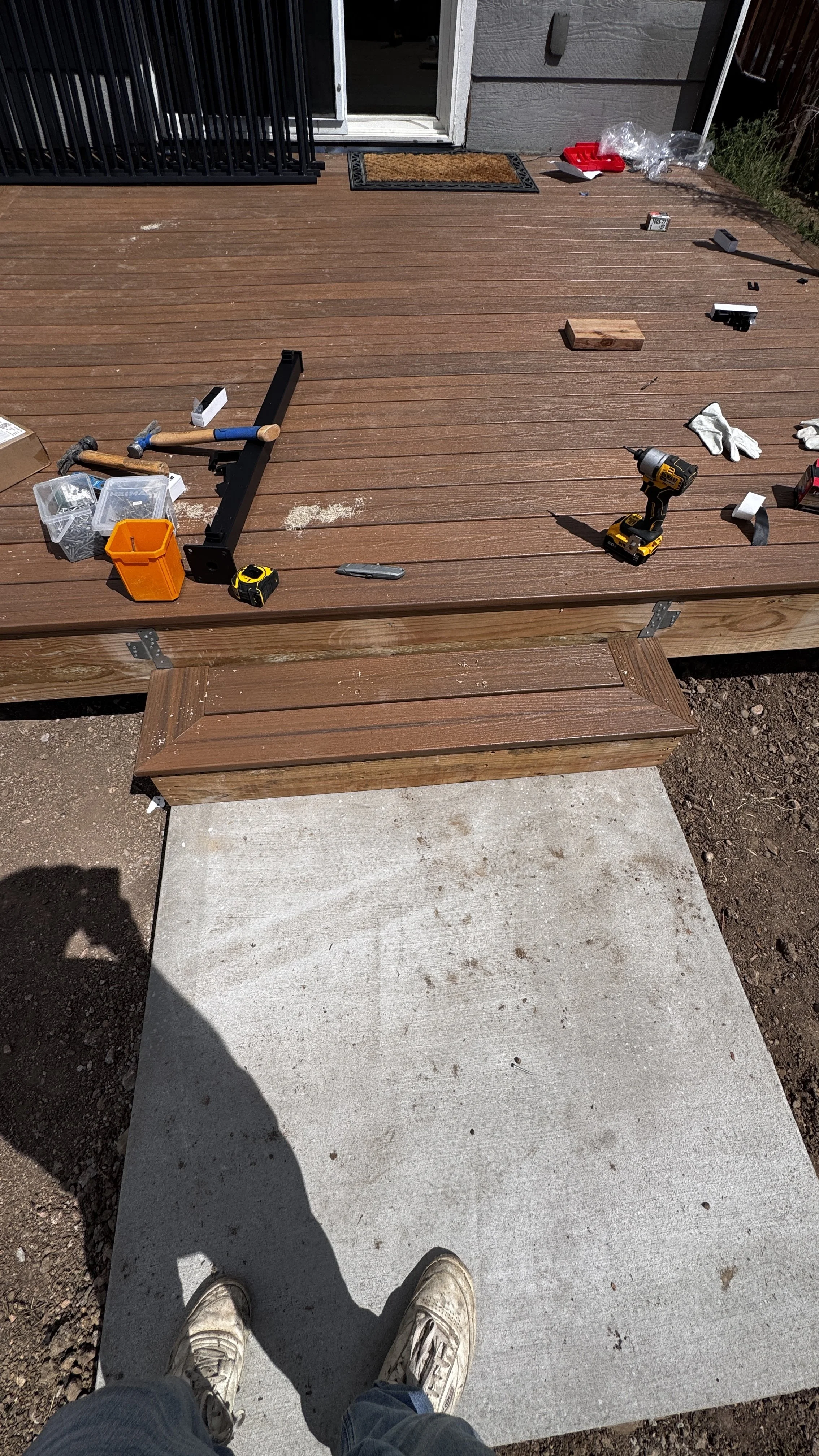 View of a newly installed wooden deck with tools and materials scattered around, with a concrete pad and shoes and shadow of person in the foreground.