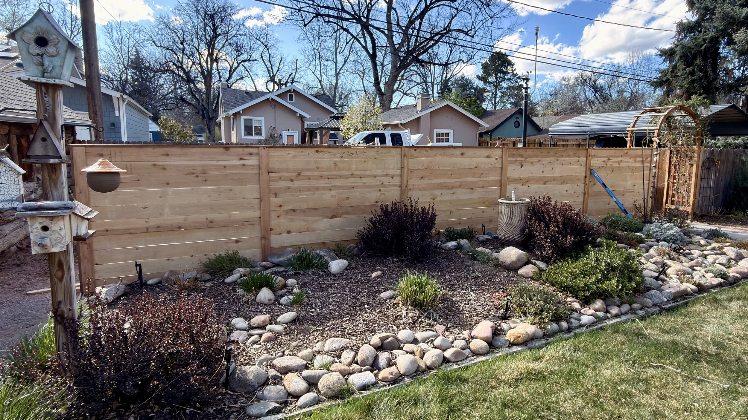 A backyard scene with a newly built wooden privacy fence, landscaped with rocks, plants, and garden features. In the background, there are neighboring houses and trees under a partly cloudy sky.