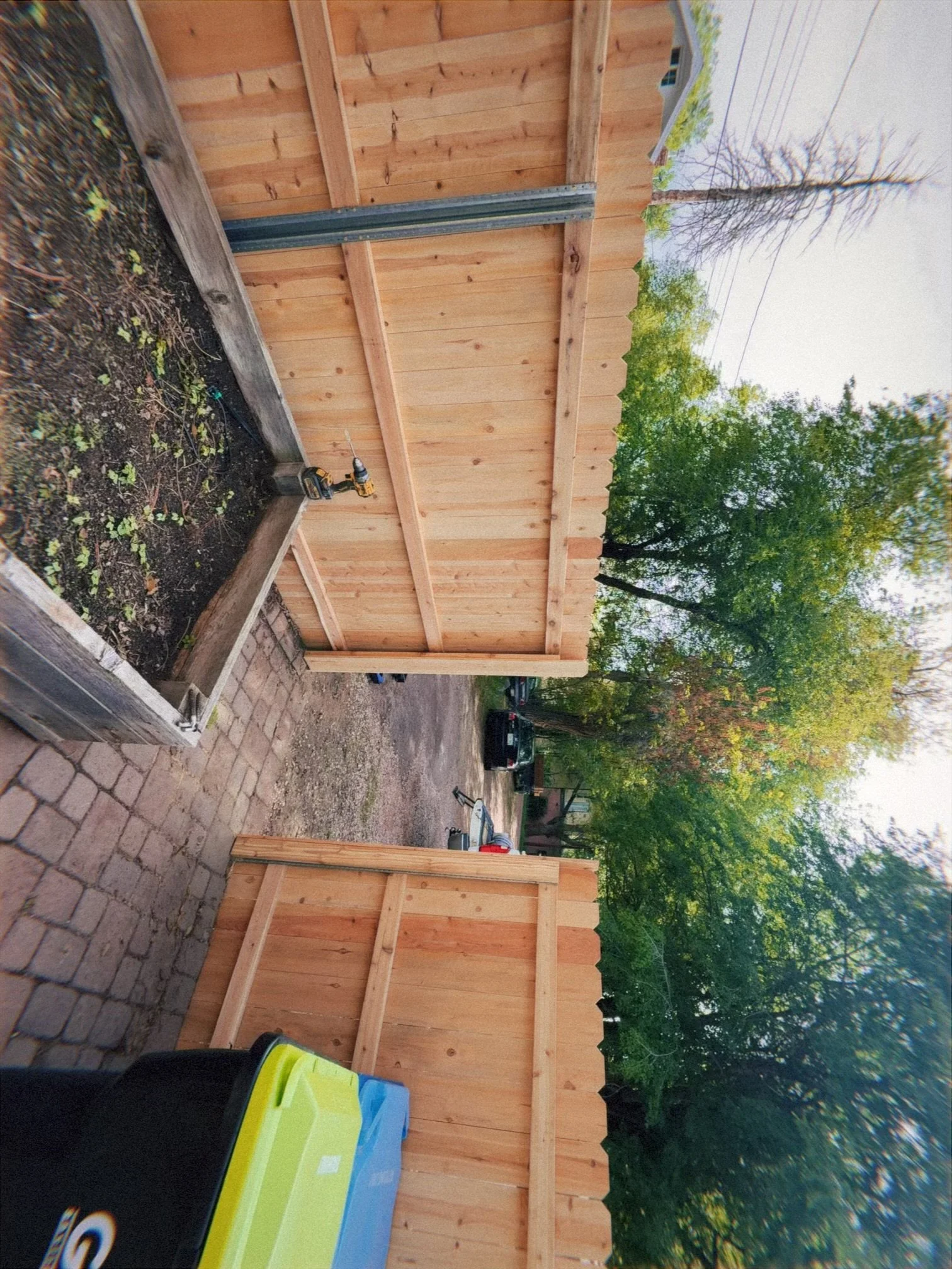 A newly built wooden fence next to a garden bed with plants, in an outdoor backyard setting with trees and a power line visible in the background.