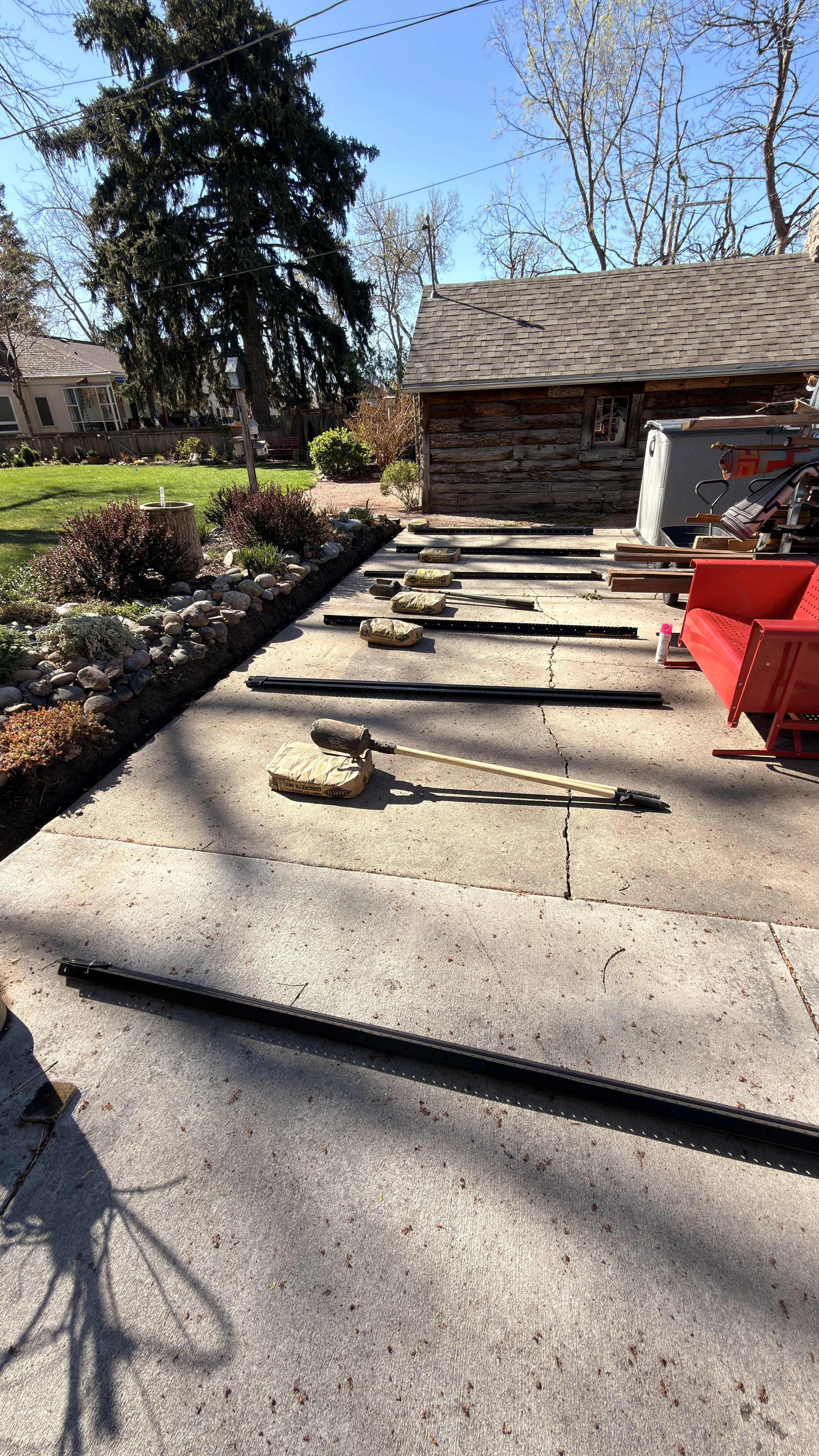 Outdoor backyard scene with gardening tools and materials on concrete, a small wooden shed, landscaped flower bed with rocks, trees, and a lawn under a clear blue sky.