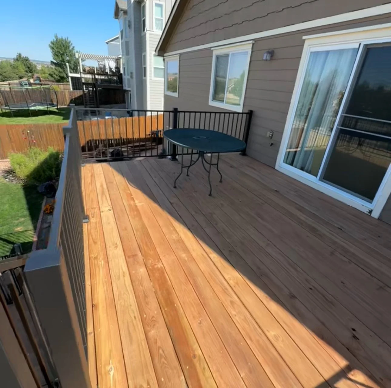 Backyard deck with wooden flooring, black metal table, and sliding glass door leading inside a house, enclosed by a wooden fence.