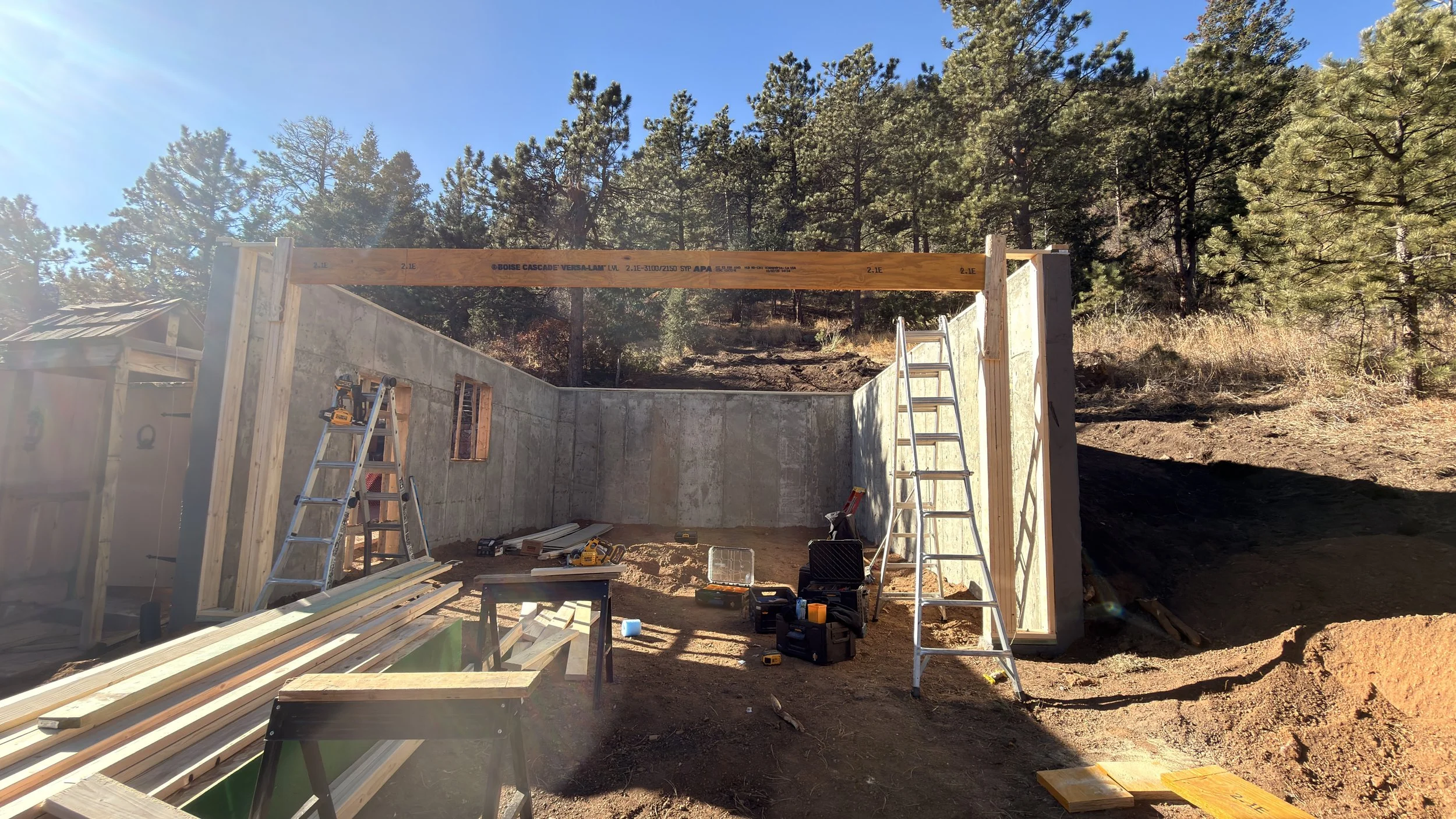 Construction site with a partially built concrete wall, wooden beams, ladders, and construction tools surrounded by trees under a clear blue sky.