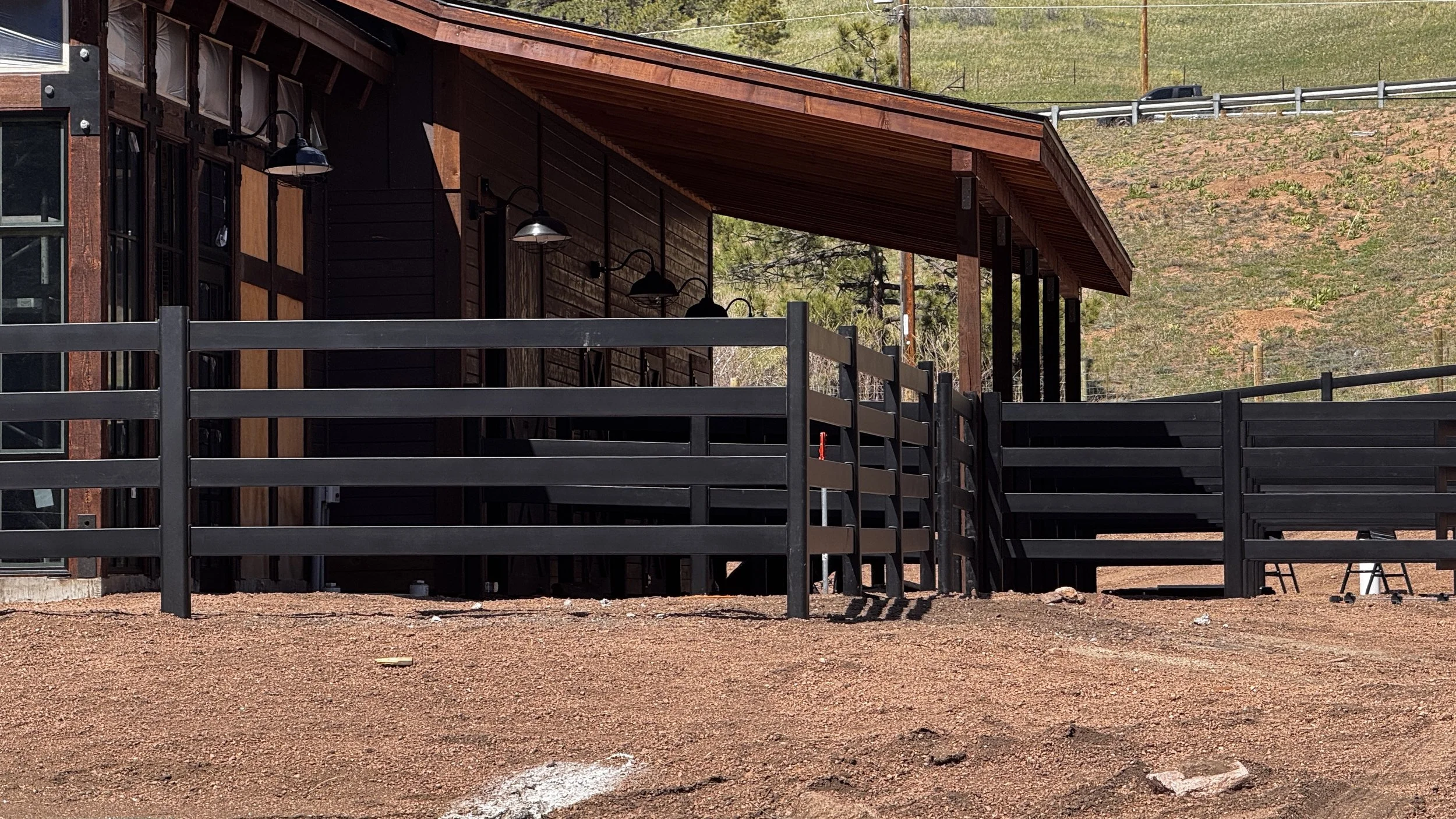 A building with a wooden exterior, black outdoor lights, and a balcony with black railing, set against a hillside with sparse vegetation.