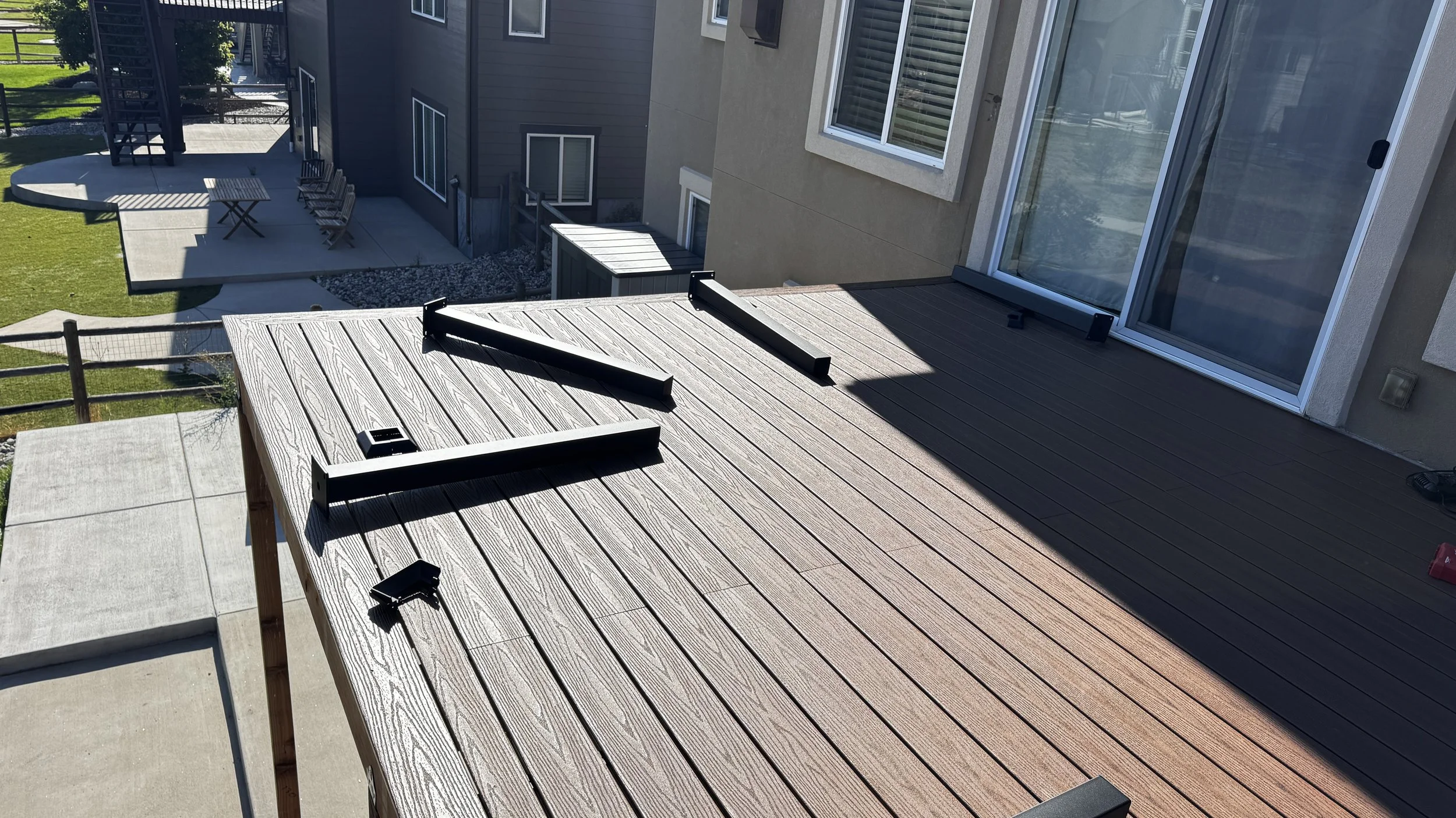 View of an outdoor composite deck with fortress handrail connected to a house with large sliding glass doors, overlooking a backyard with a patio area and grass lawn, with furniture and shadows indicating a sunny day.