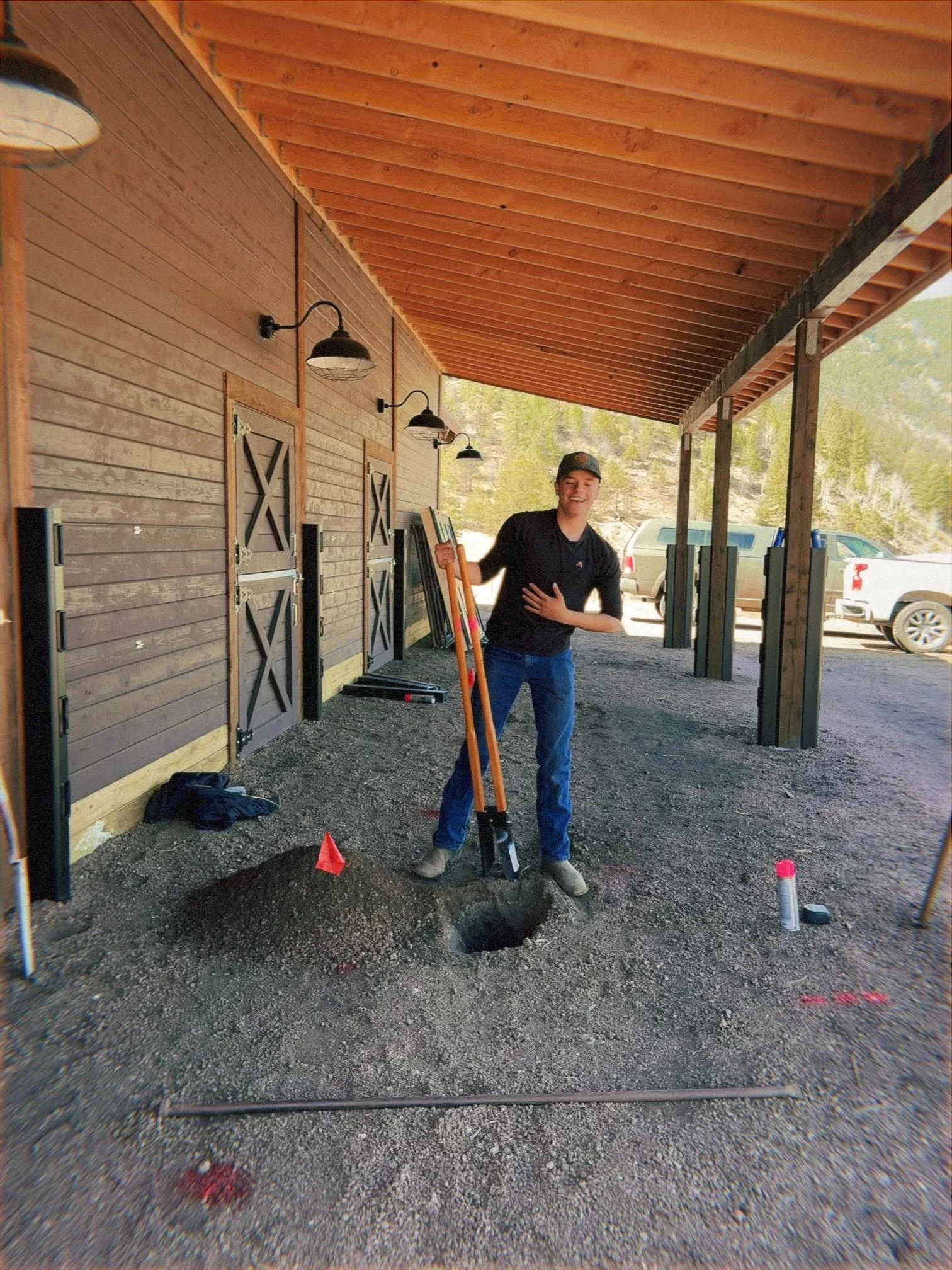 Young man smiling and holding surveying equipment on a gravel driveway outside a wooden building with a porch and parked vehicles in the background.