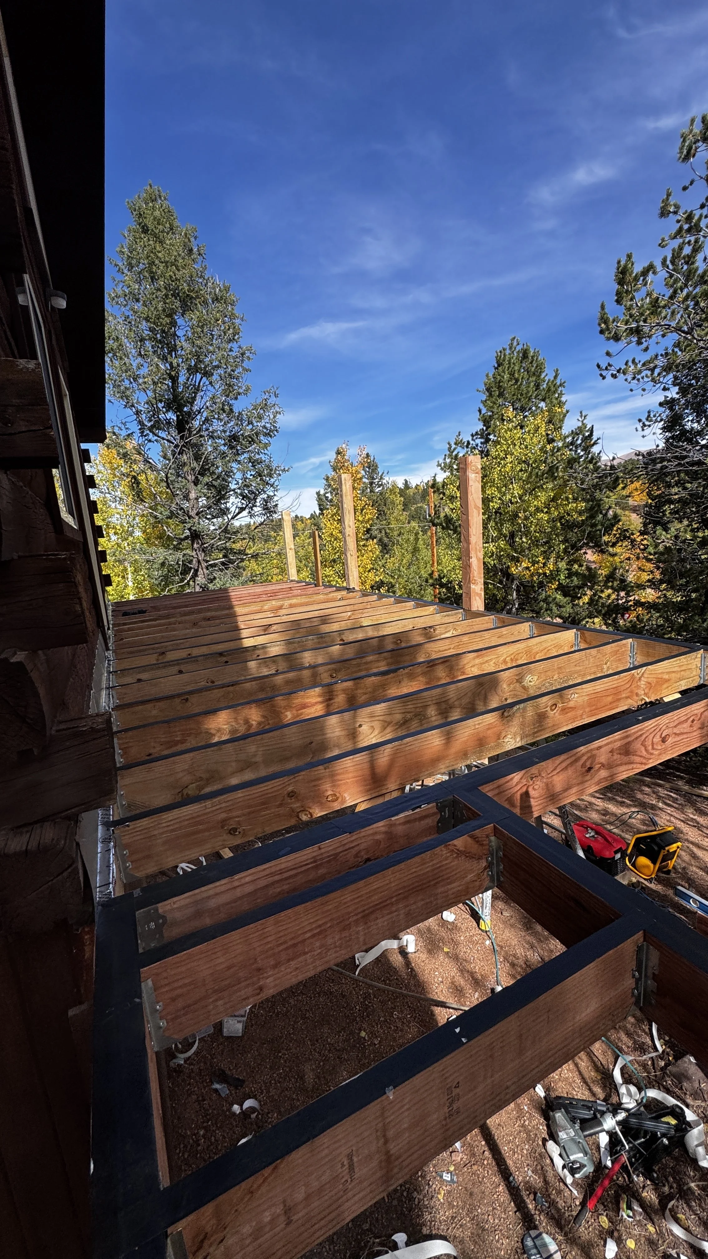 Construction scene on a wooden deck with support beams, tools, and equipment, with trees and a blue sky in the background.