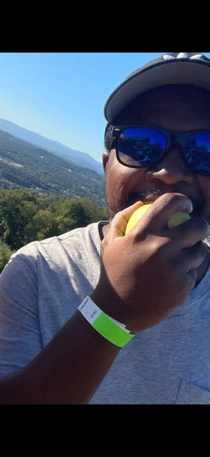 A man wearing sunglasses and a cap, eating a yellow fruit outdoors with a scenic view of trees and mountains in the background.