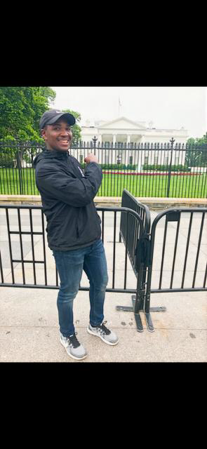 A young man standing outside near a fence in front of the White House, smiling and pointing at it.