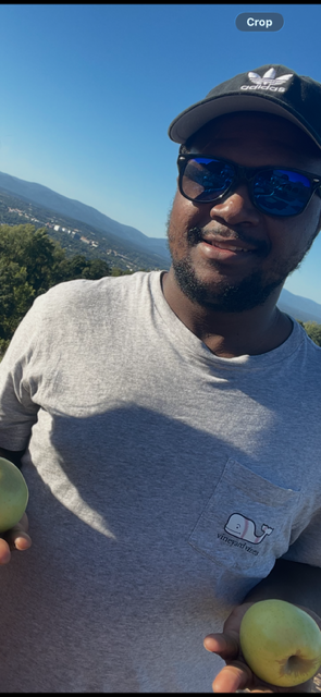 A man wearing a gray t-shirt, a black cap, and sunglasses holding two apples outdoors with a landscape and mountains in the background.