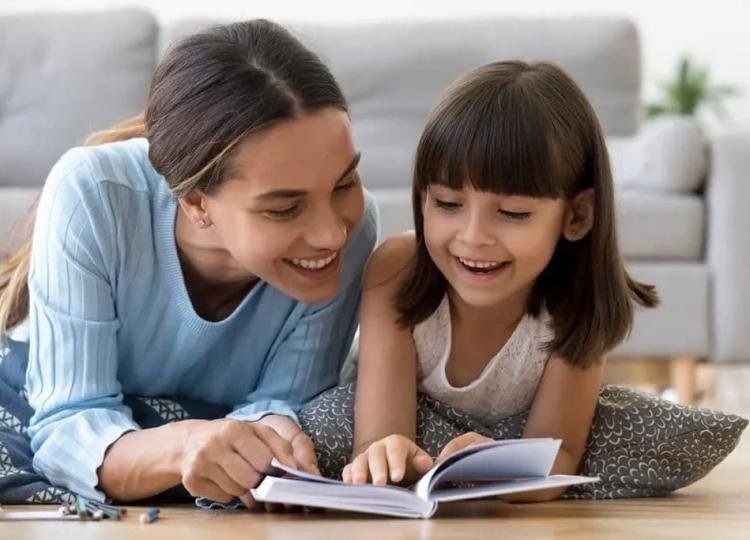 A woman and a young girl are lying on the floor together, reading a book and smiling.