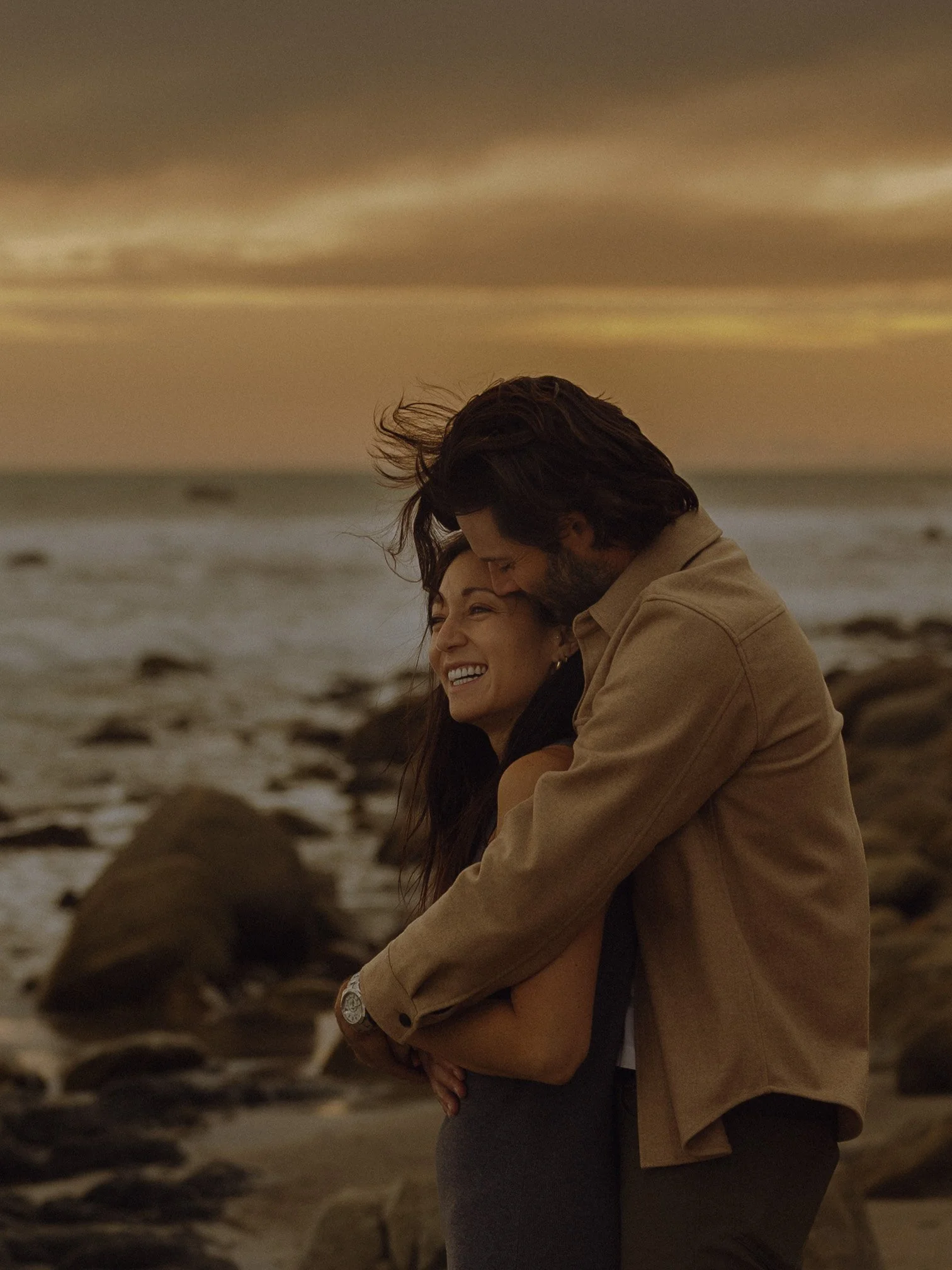 A couple hugging on the beach during sunset, with rocks and ocean in the background.