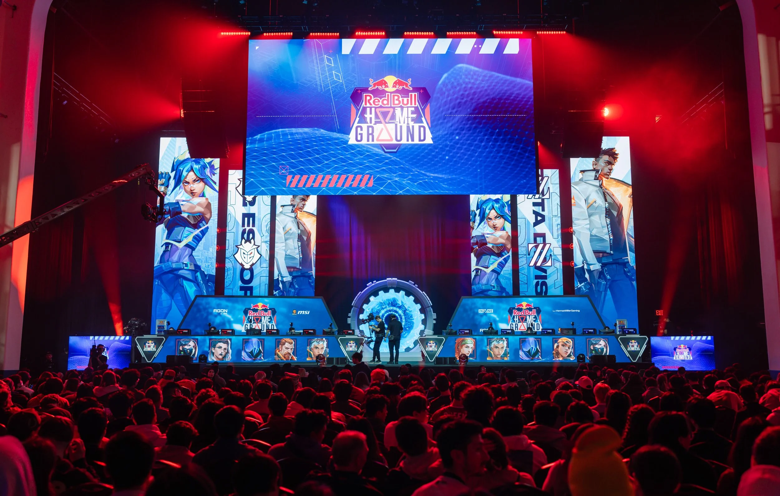 A large esports event stage with a crowd in front. The stage features a central big screen with the 'Red Bull Home Ground' logo and characters from a video game. Flanking the main screen are smaller screens displaying animated characters, and the audience is seated and watching the competition.