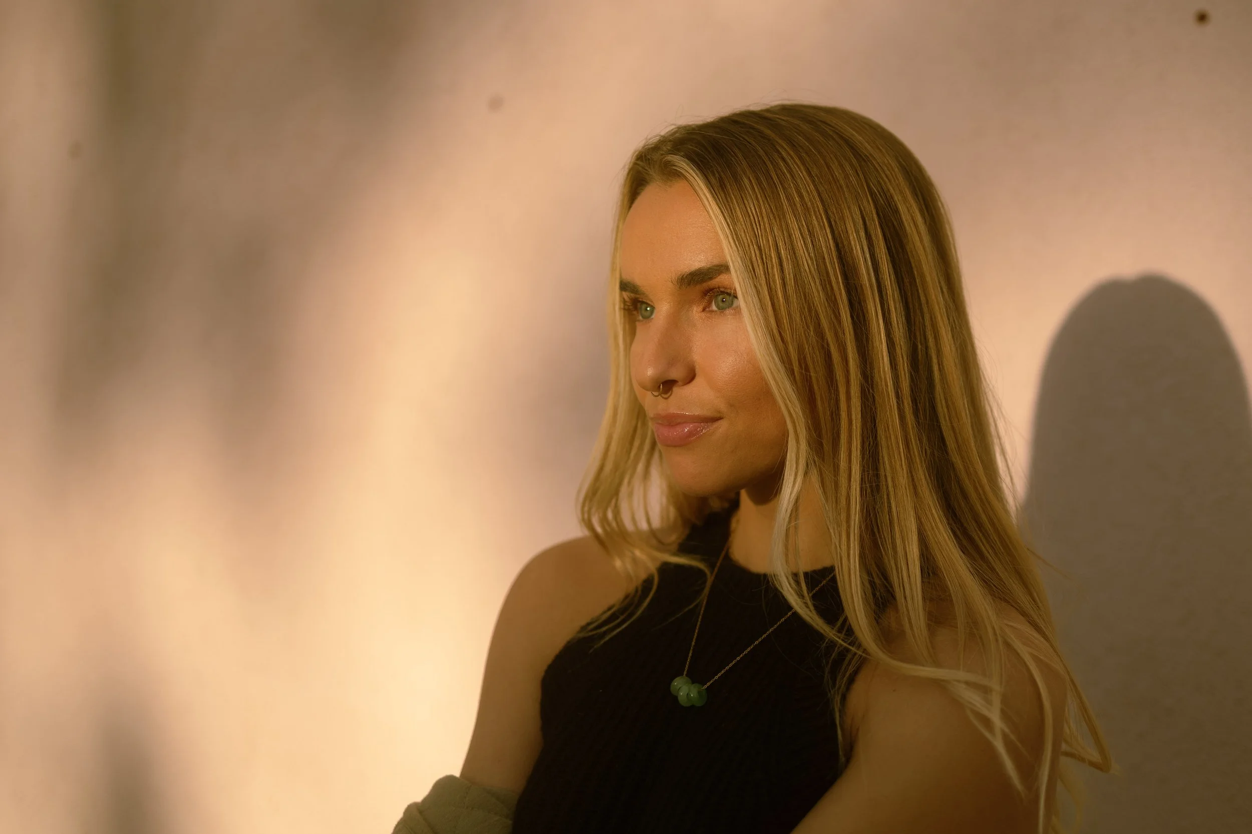 A young woman with long blonde hair, green eyes, and a septum piercing, wearing a black sleeveless top and a green beaded necklace, is smiling softly while looking to her right. The background is softly lit and blurred, with warm tones.