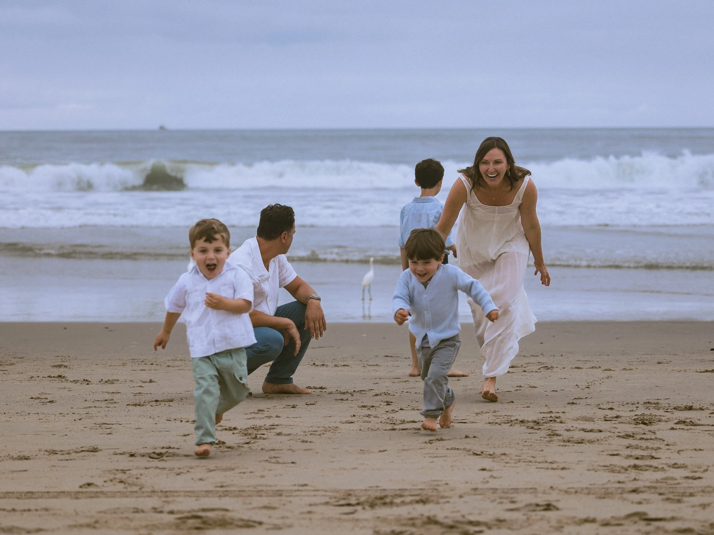 A woman and several children are playing and running on a sandy beach near the ocean, with waves in the background and seagulls on the sand.