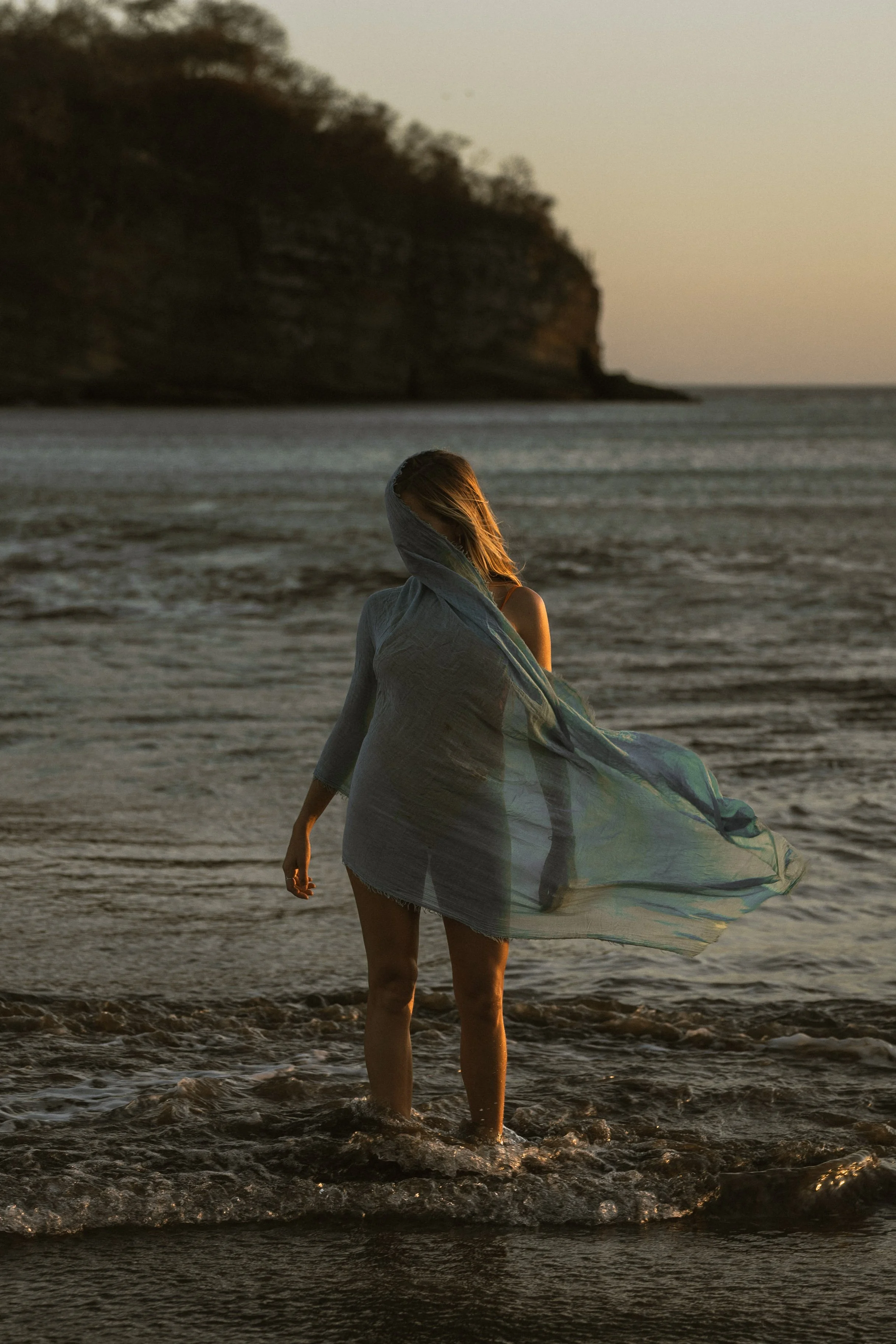 A woman with blonde hair wrapped in a light-colored cloth, standing in the shallow waters of a beach at sunset, with a rocky cliff in the background.
