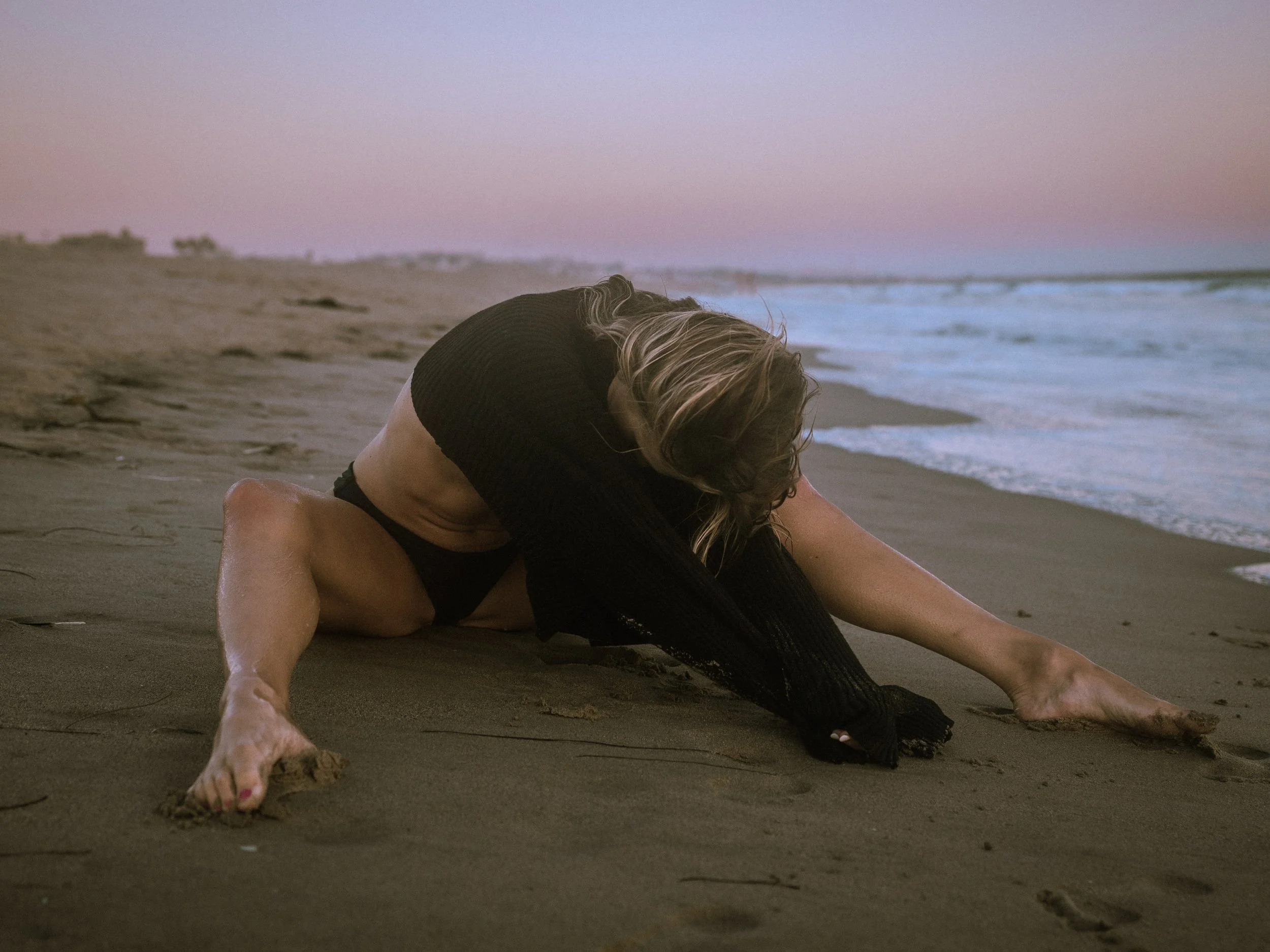 A person with blonde hair doing a backbend stretch on the sandy beach at sunset with the ocean waves in the background.