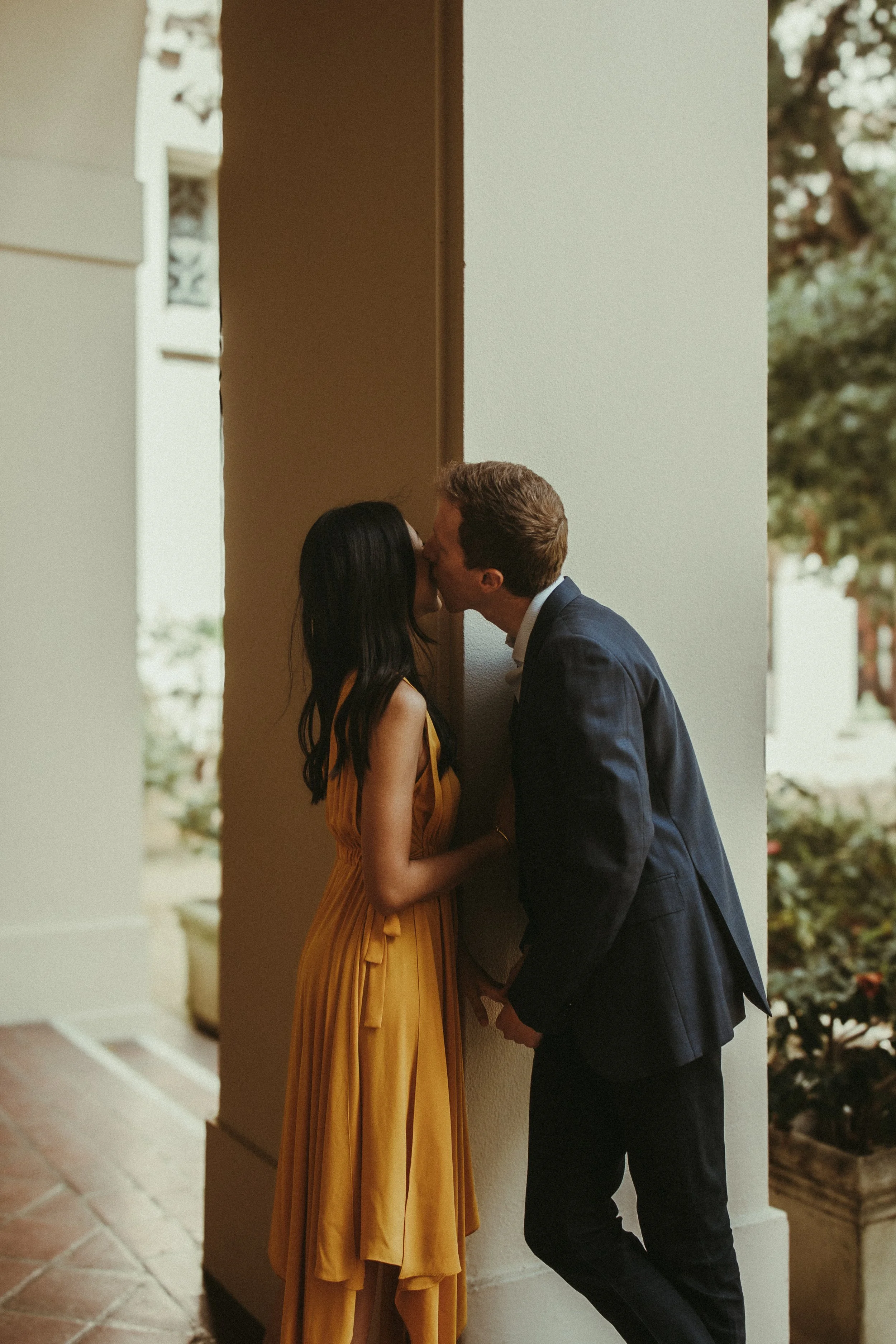 A couple sharing a kiss under a covered outdoor area, with the woman wearing a yellow dress and the man in a dark suit.