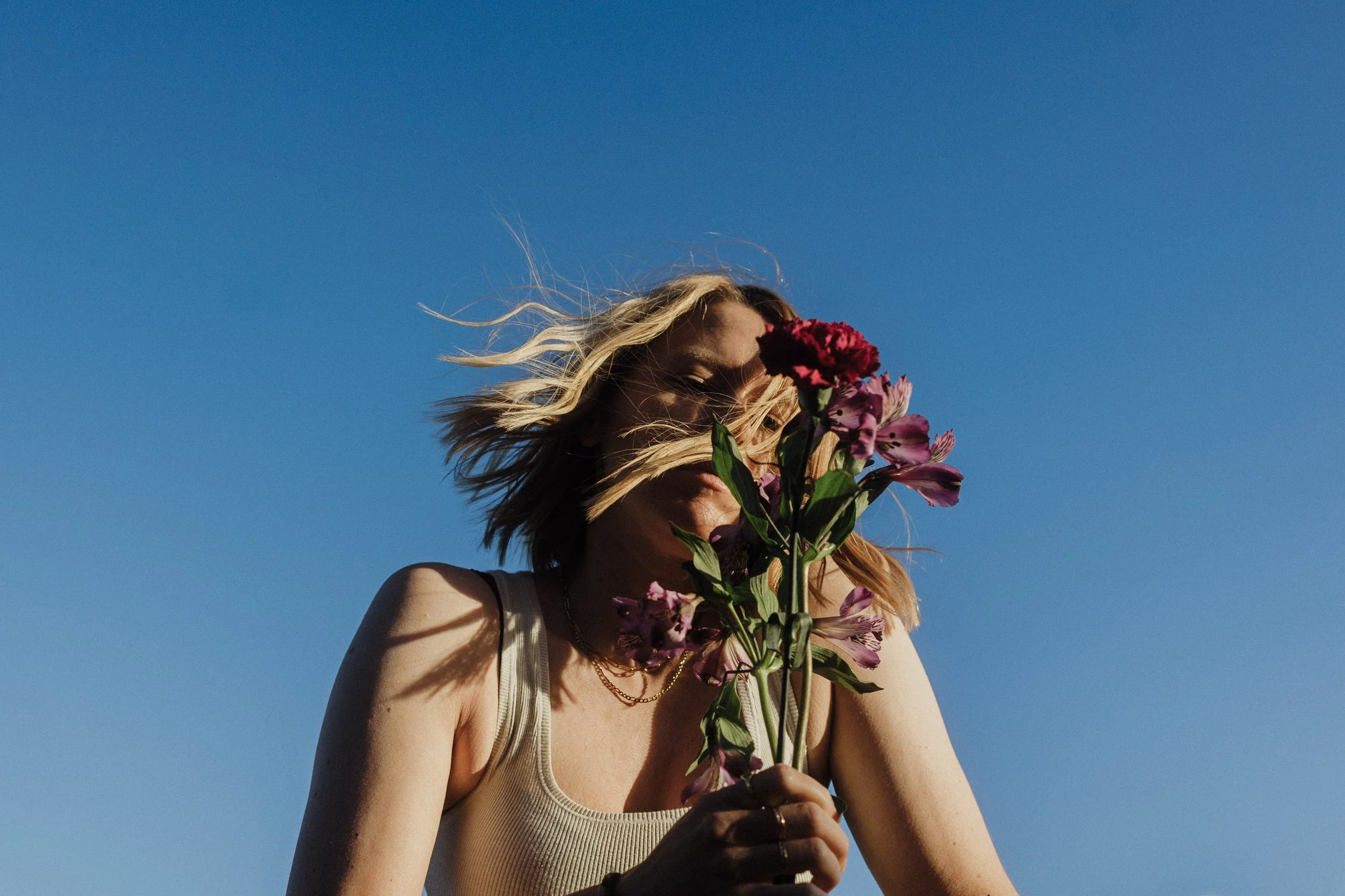 A woman with blonde hair holding a bouquet of purple and pink flowers in front of her face, standing against a clear blue sky.