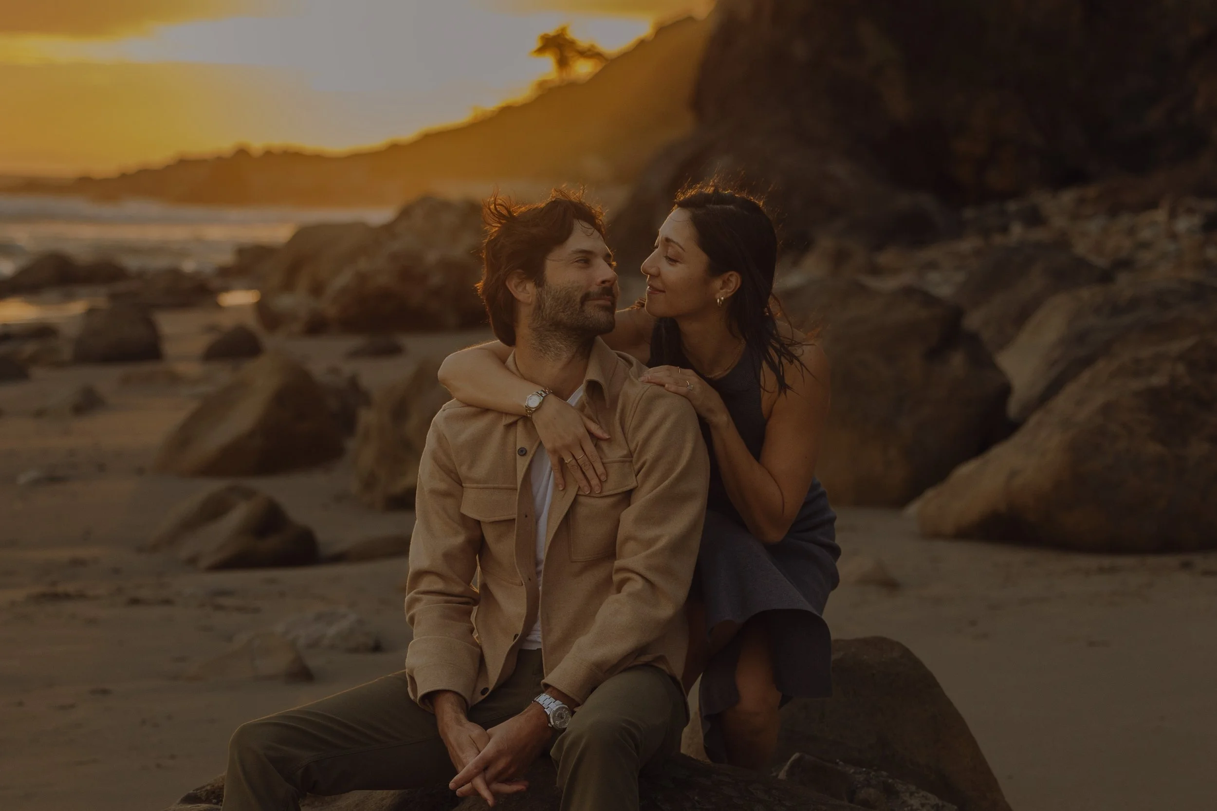 A man and woman sitting on a rocky beach during sunset, with the woman leaning close to the man's face, both smiling and enjoying each other's company.