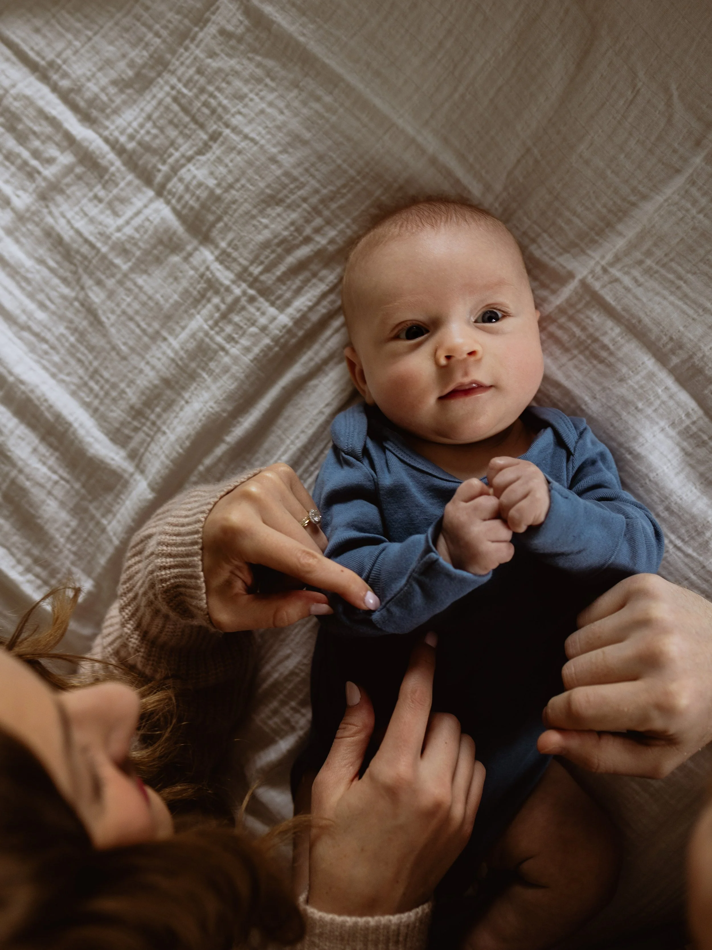 A baby with light skin and light-colored hair lying on a bed with a beige quilted blanket. The baby is wearing a blue onesie and has their hands clasped together. Several adults’ hands are gently touching the baby, including one hand with a ring.