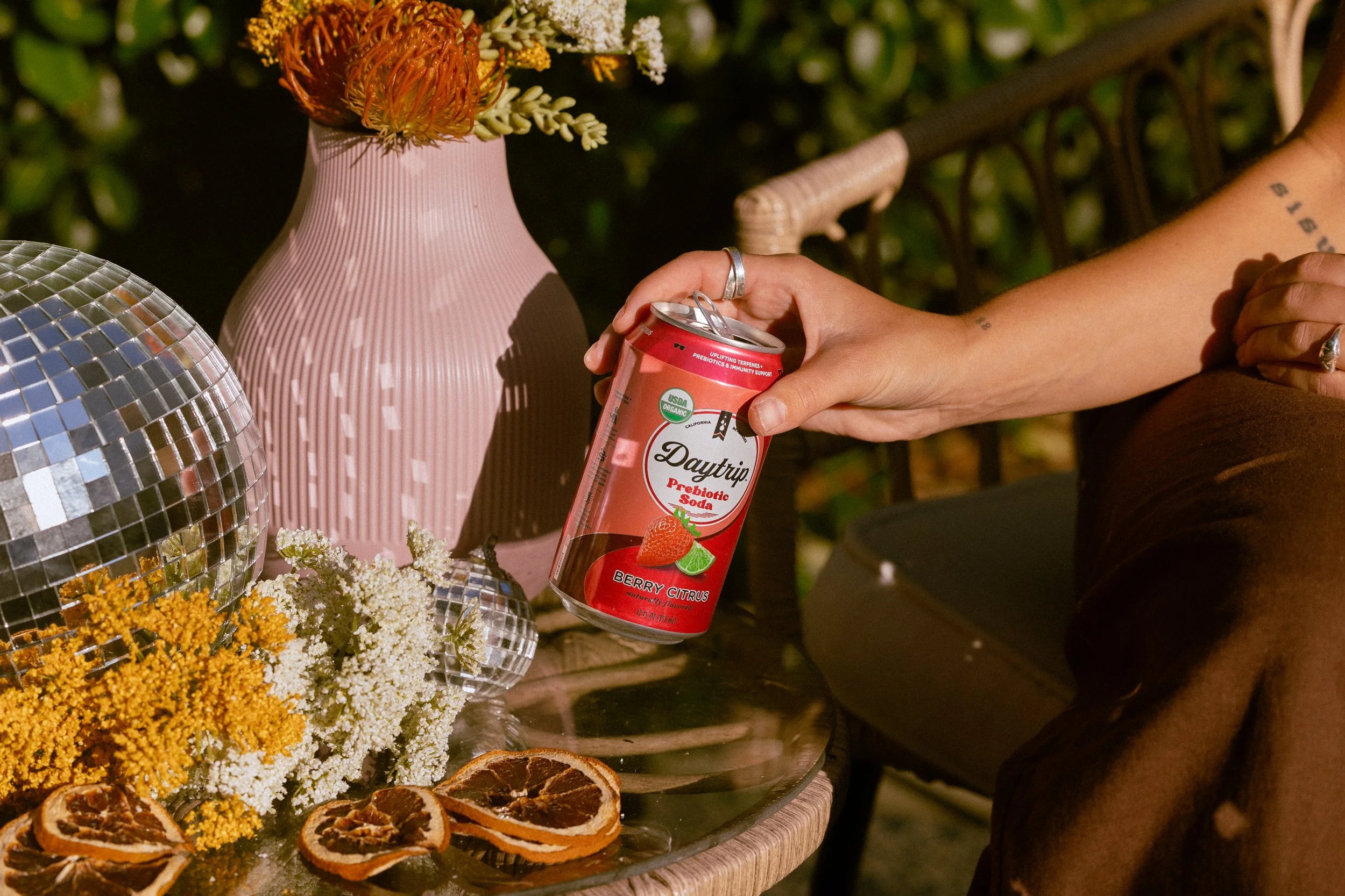 Person holding a can of Daytrip prebiotic soda near floral arrangements and dried citrus slices on a table outdoors at night.