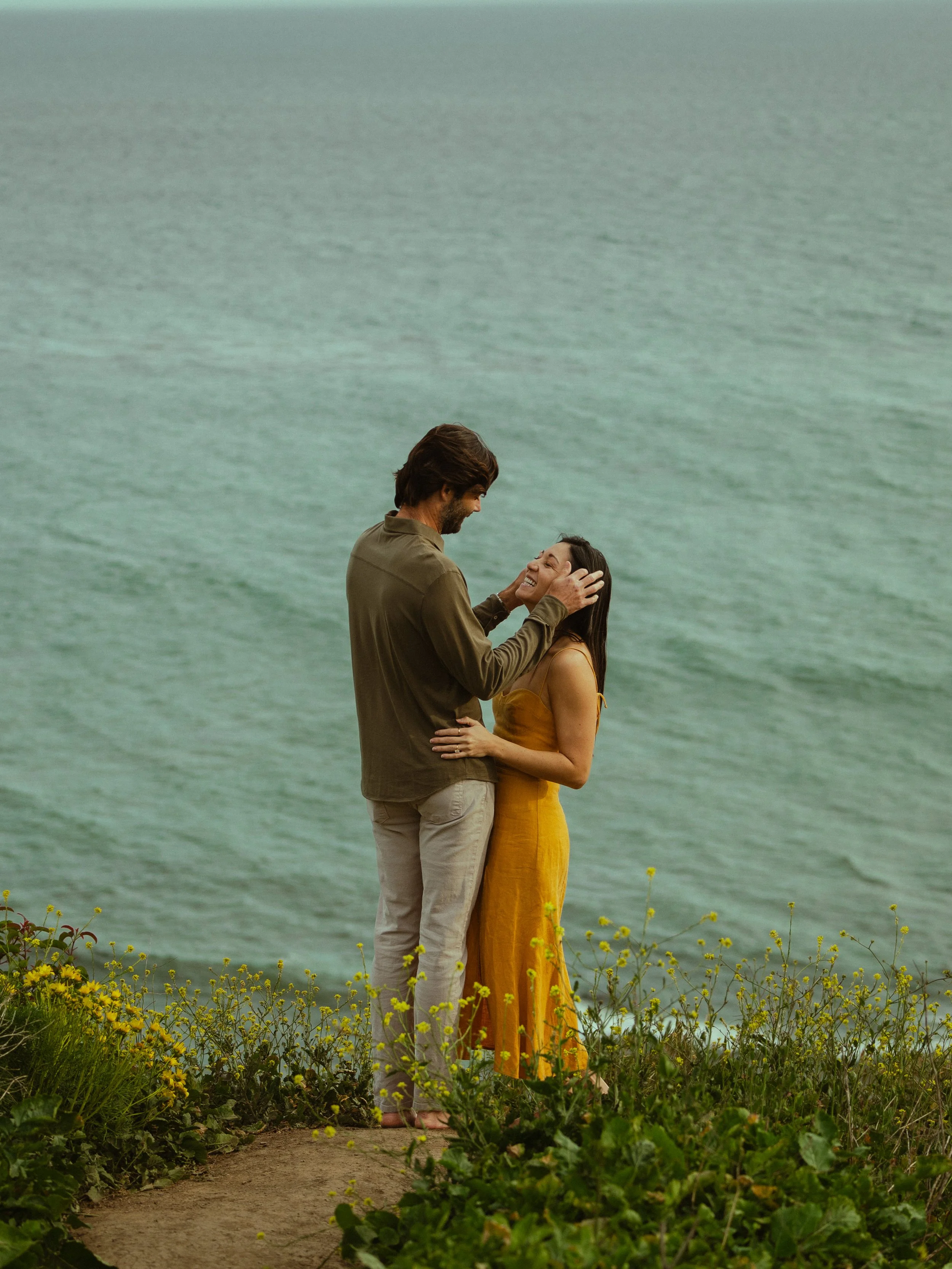A man and woman standing close together on a garden path near the water, with yellow flowers surrounding them, sharing an intimate moment and smiling.