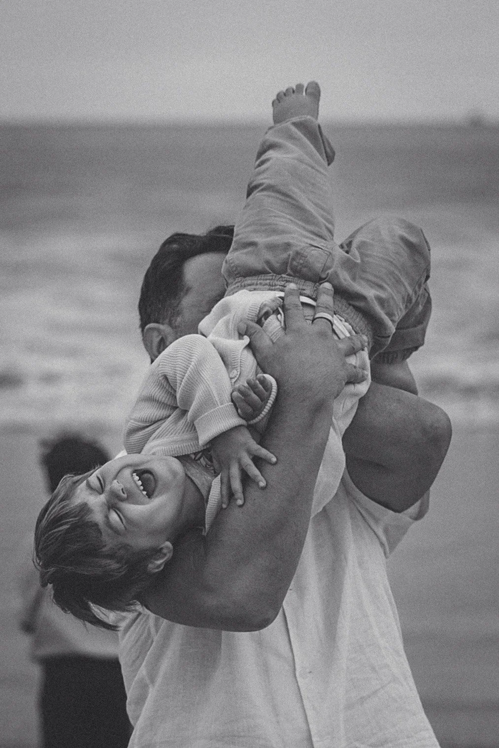 A man holding a laughing boy upside down at the beach, with ocean waves in the background.