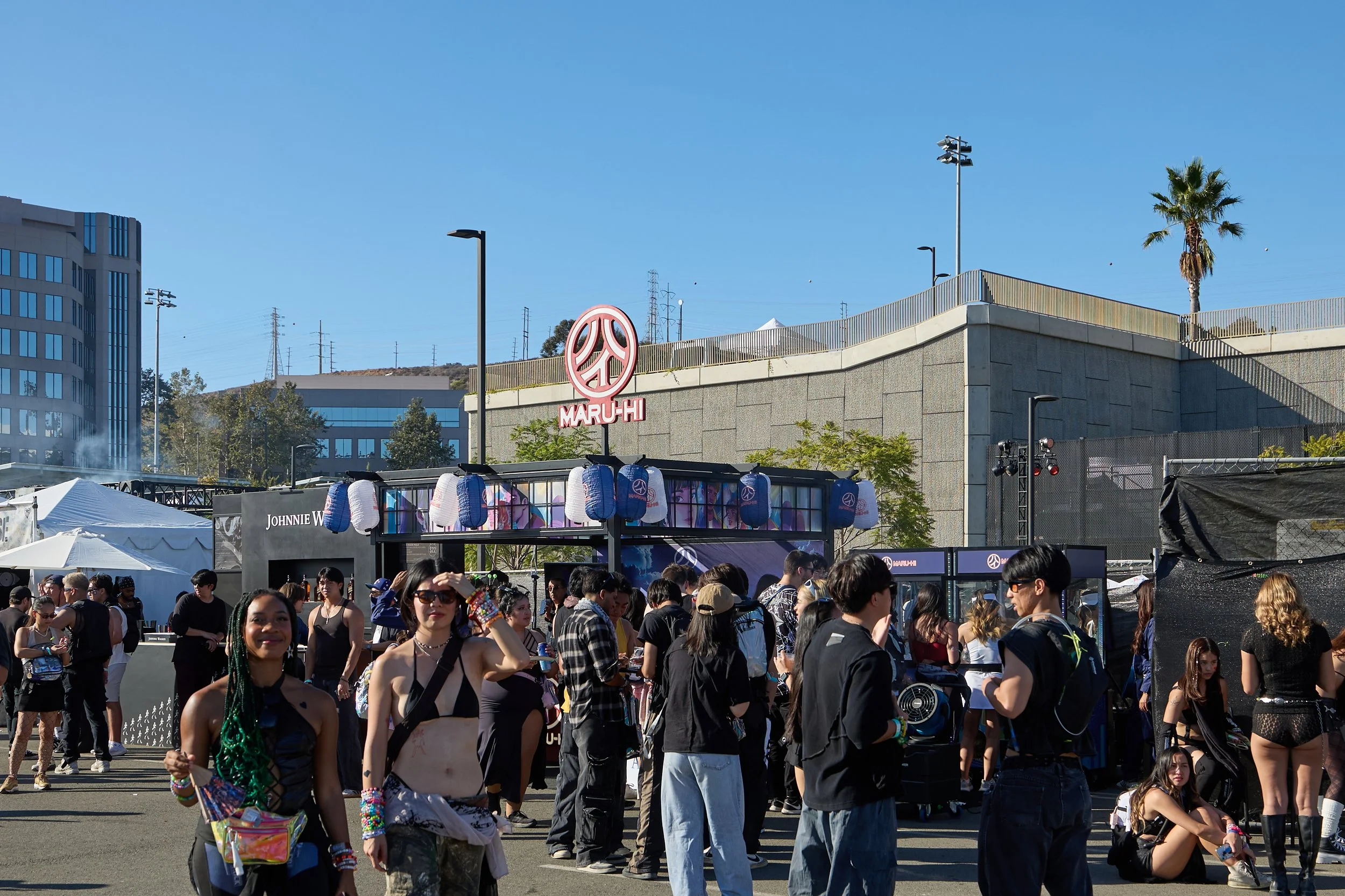 Crowd of people gathered outdoors at a festival or event, with tents and stage setup in the background, including a Maru-Hi sign and blue sky.