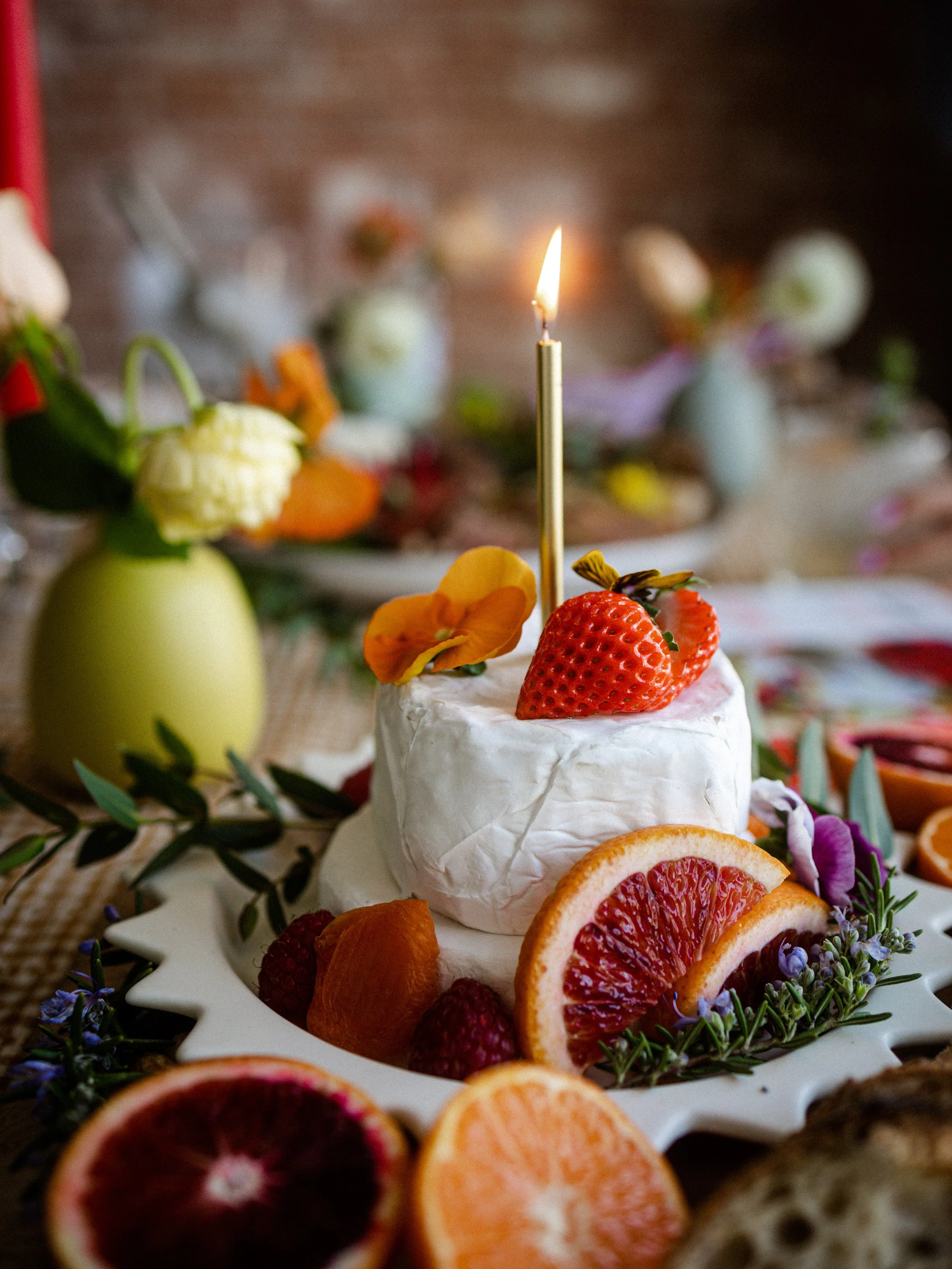 A decorated table with a cake topped with strawberries and a lit candle, surrounded by sliced citrus fruits and flowers.