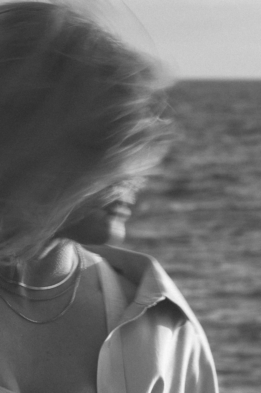 A black and white photo of a woman with long hair, wearing a collared shirt and layered necklaces, looking down by the water.