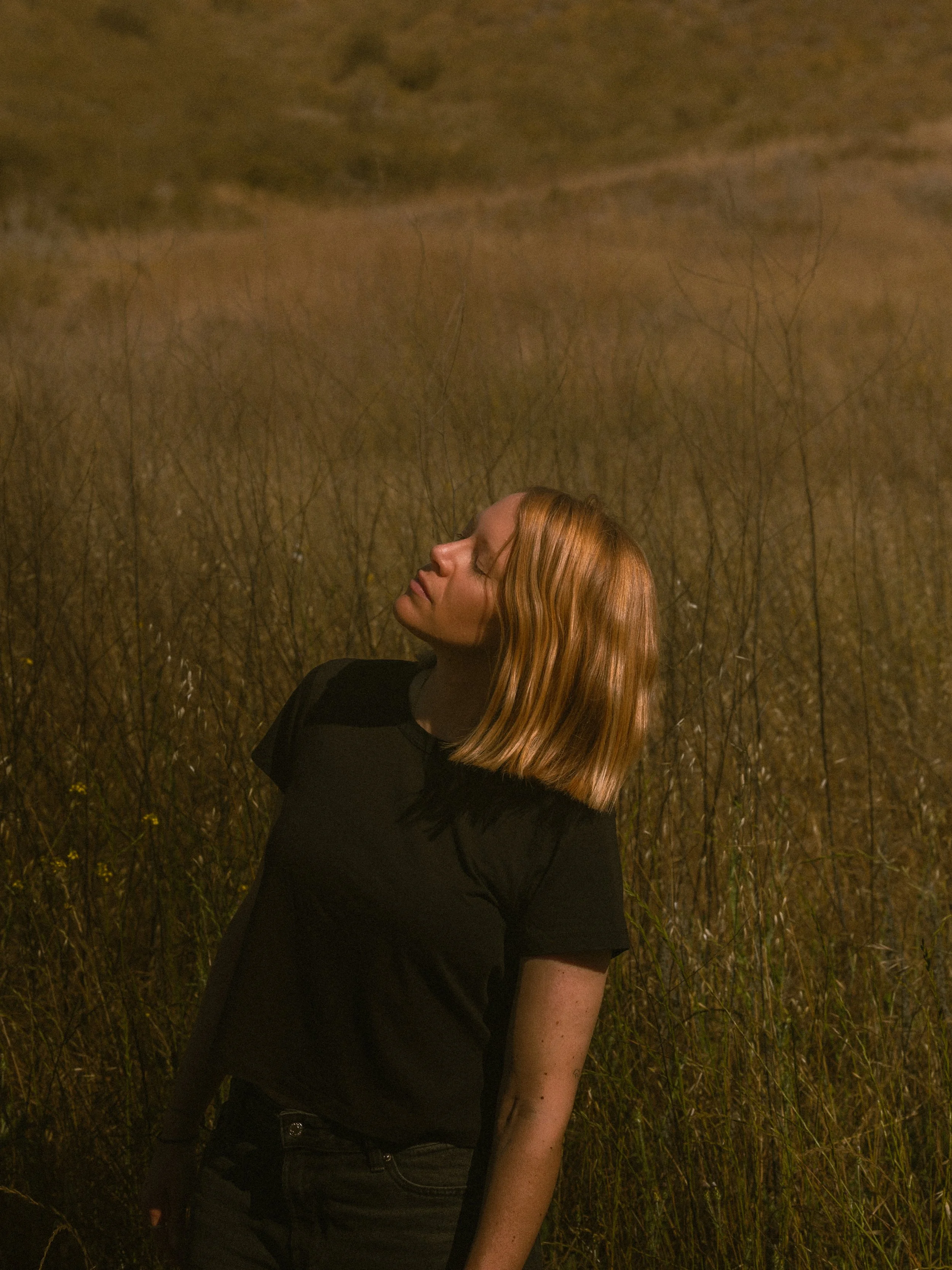 A woman with red hair, closed eyes, and a black shirt standing in a field of tall, dry grass at night, with a dark sky in the background.
