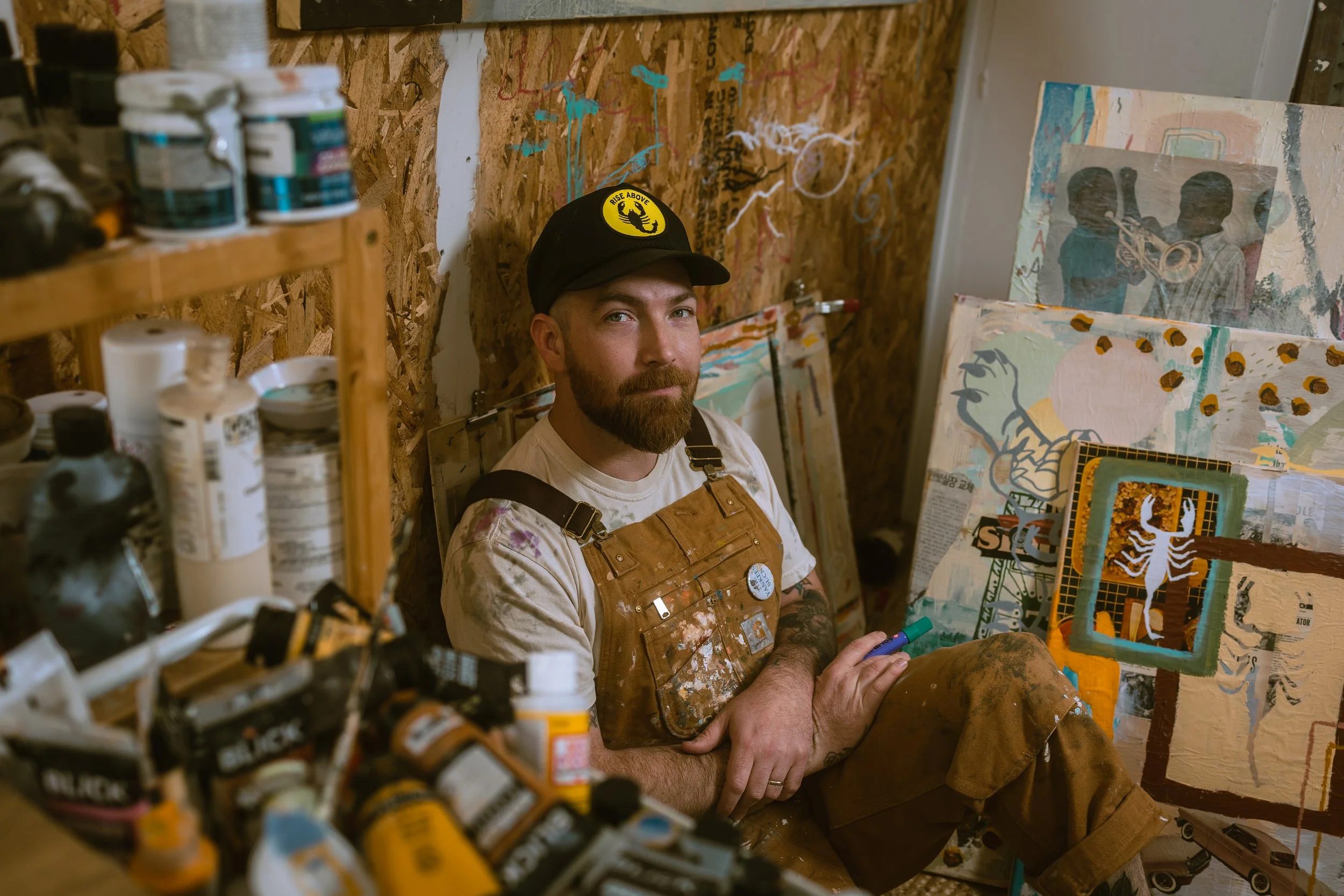 A man with a beard wearing a black cap with a yellow logo, a tie-dye shirt, and brown overalls sitting in an art studio surrounded by paintings and art supplies. He is looking at the camera with a slight smile.