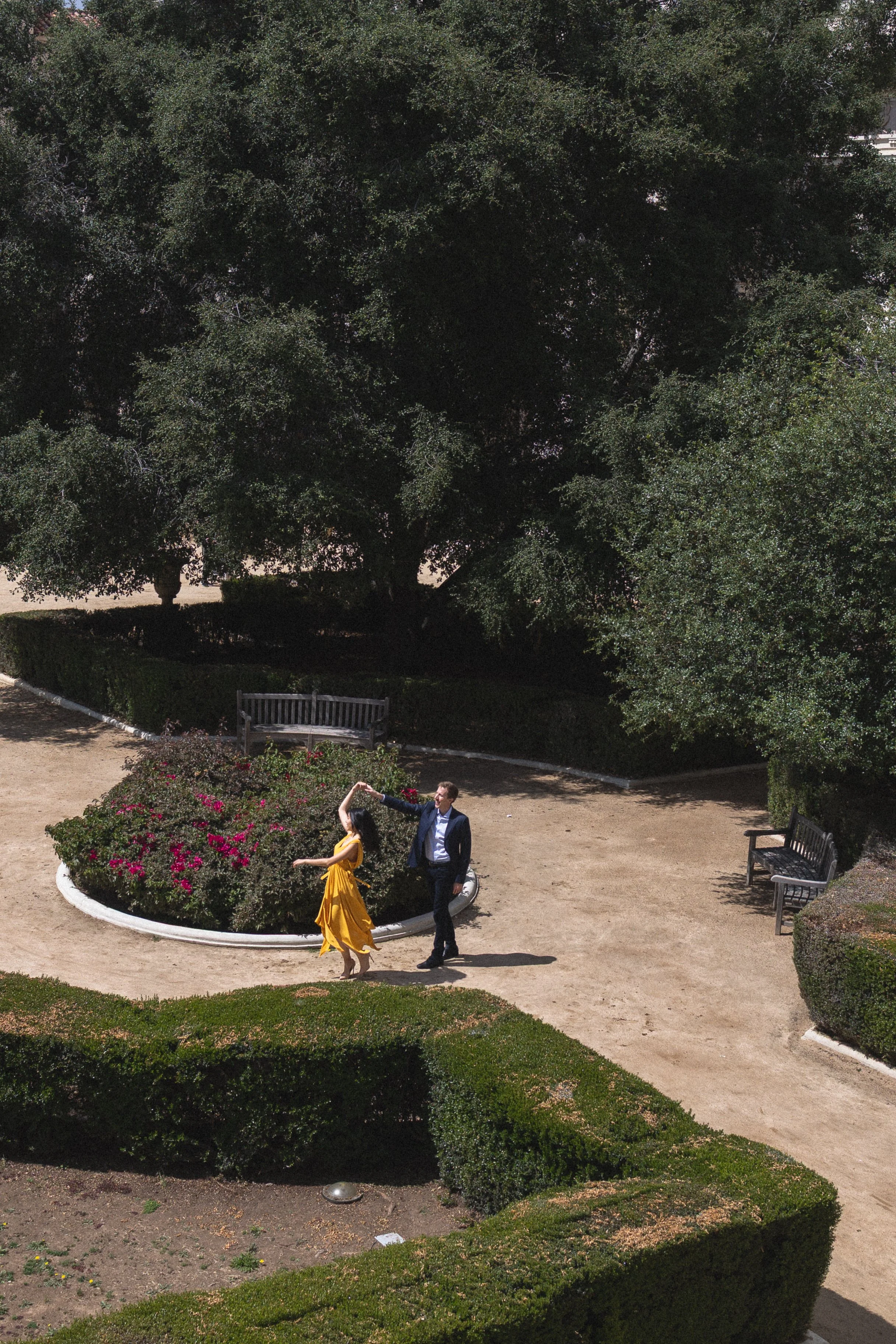A young man in a suit and a young woman in a yellow dress dancing and posing together in a garden with a flower bed and benches, shaded by large trees.