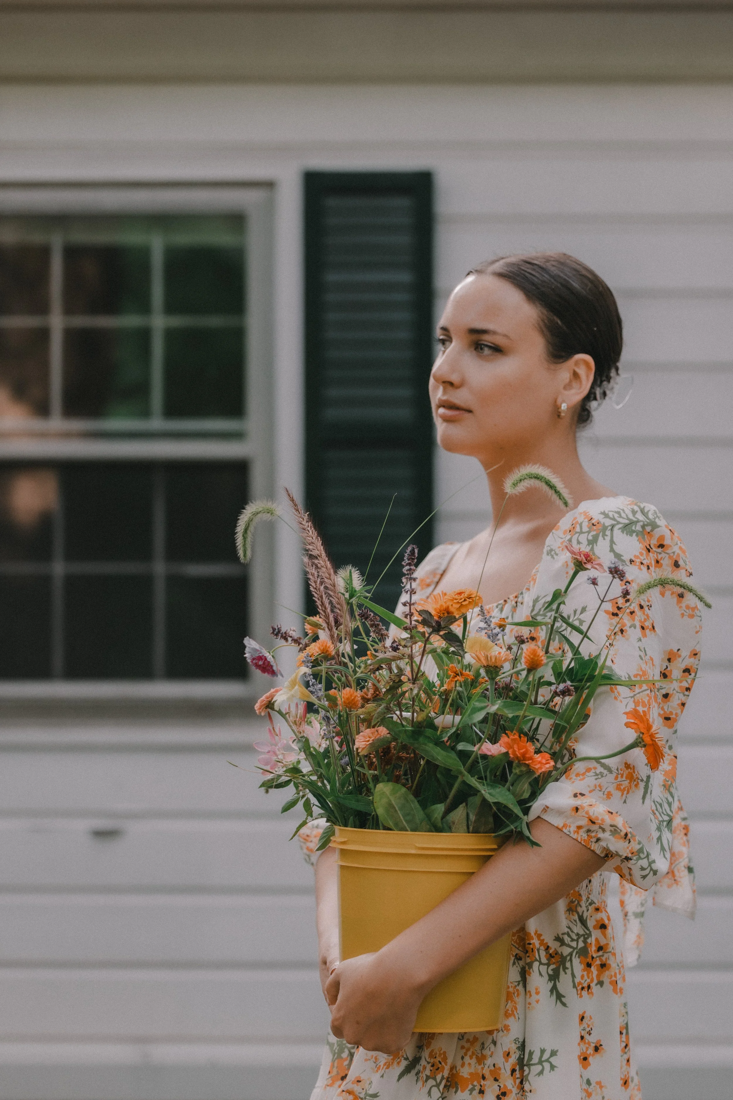 A woman with dark hair and earrings, wearing a floral dress, holding a yellow bucket filled with various flowers in front of a white house with black shutters.