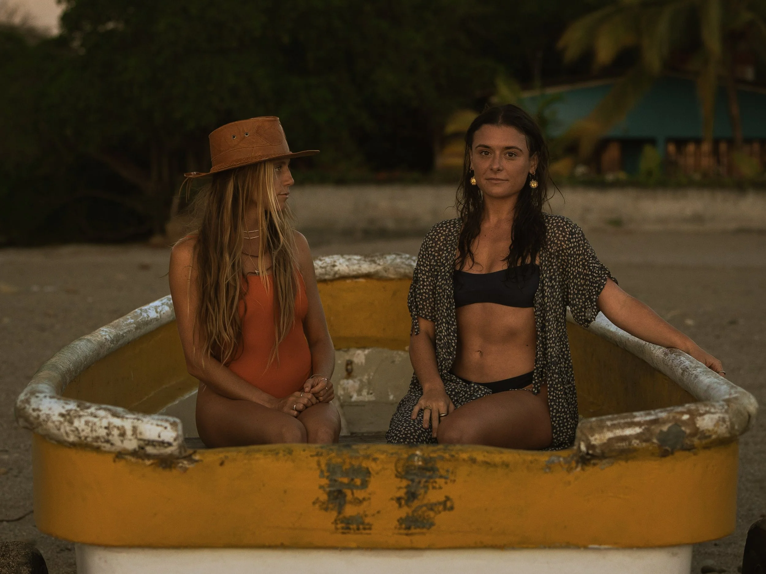 Two women sitting in a yellow boat on a beach or shoreline during sunset or dusk, with tropical trees and a hut in the background.