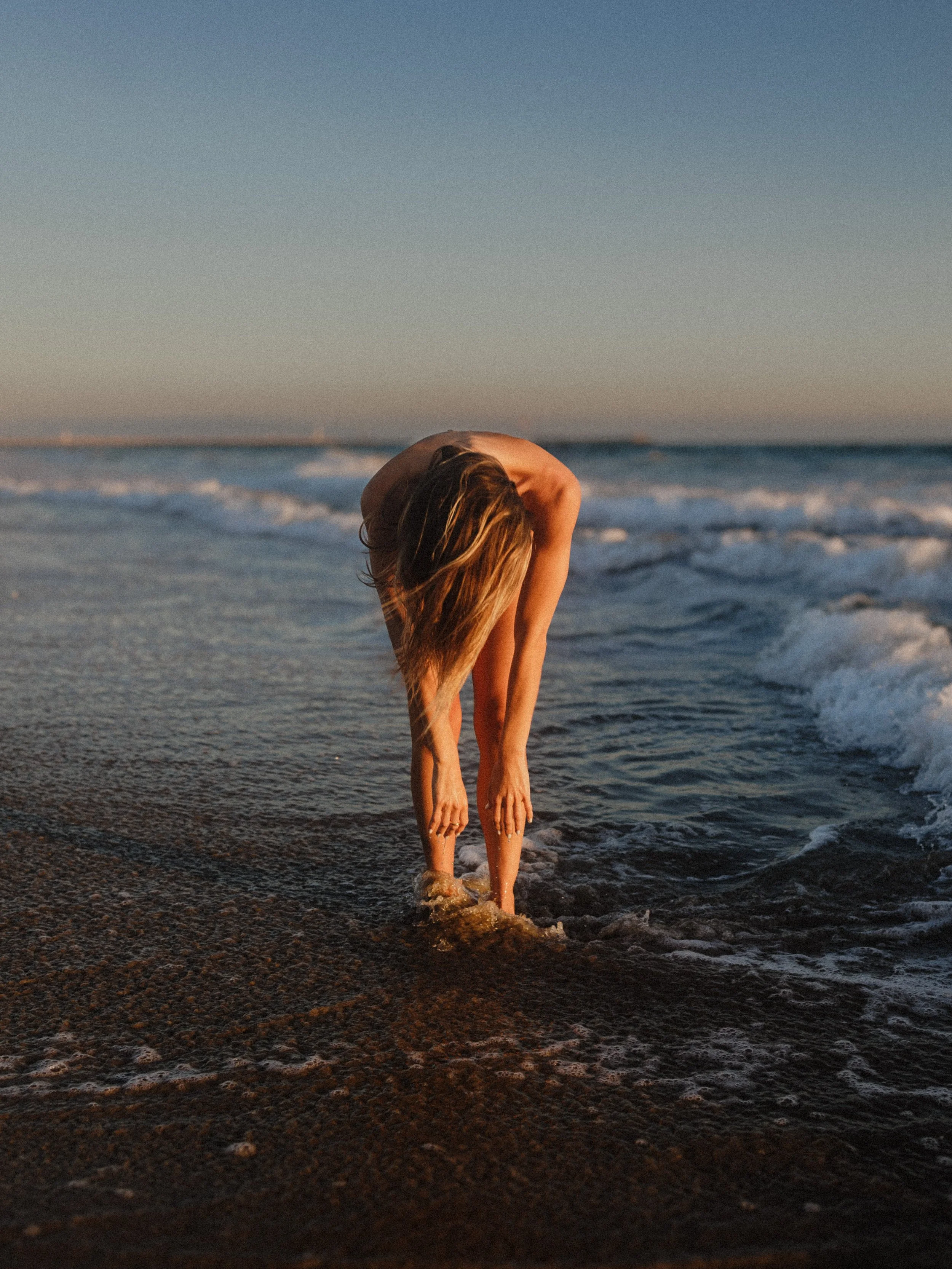 A person with long hair bends over in shallow ocean water near the shore at sunset.