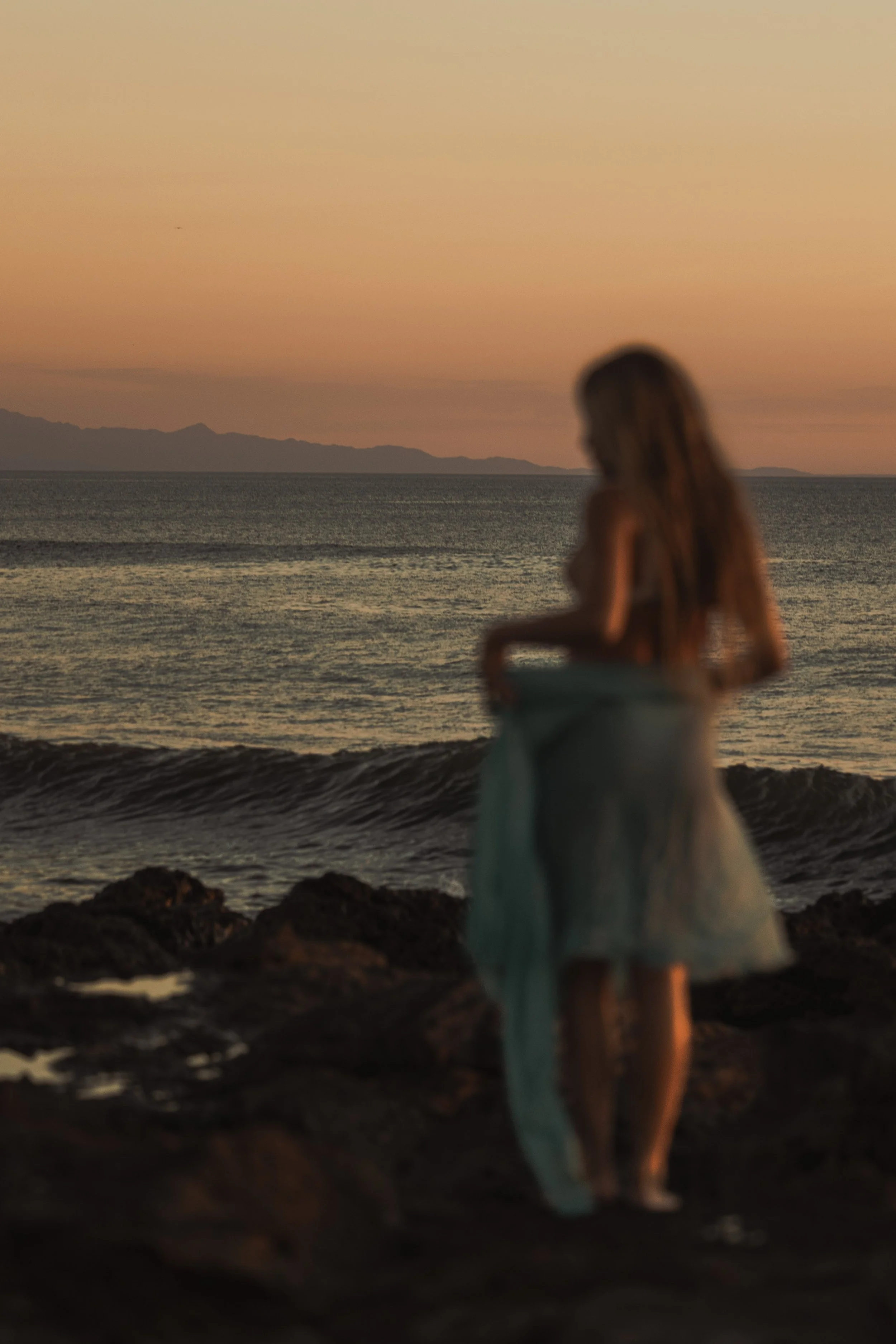 A blurry woman with long hair stands on a rocky beach at sunset, looking at the ocean, with mountains in the background and the sky painted in shades of orange.