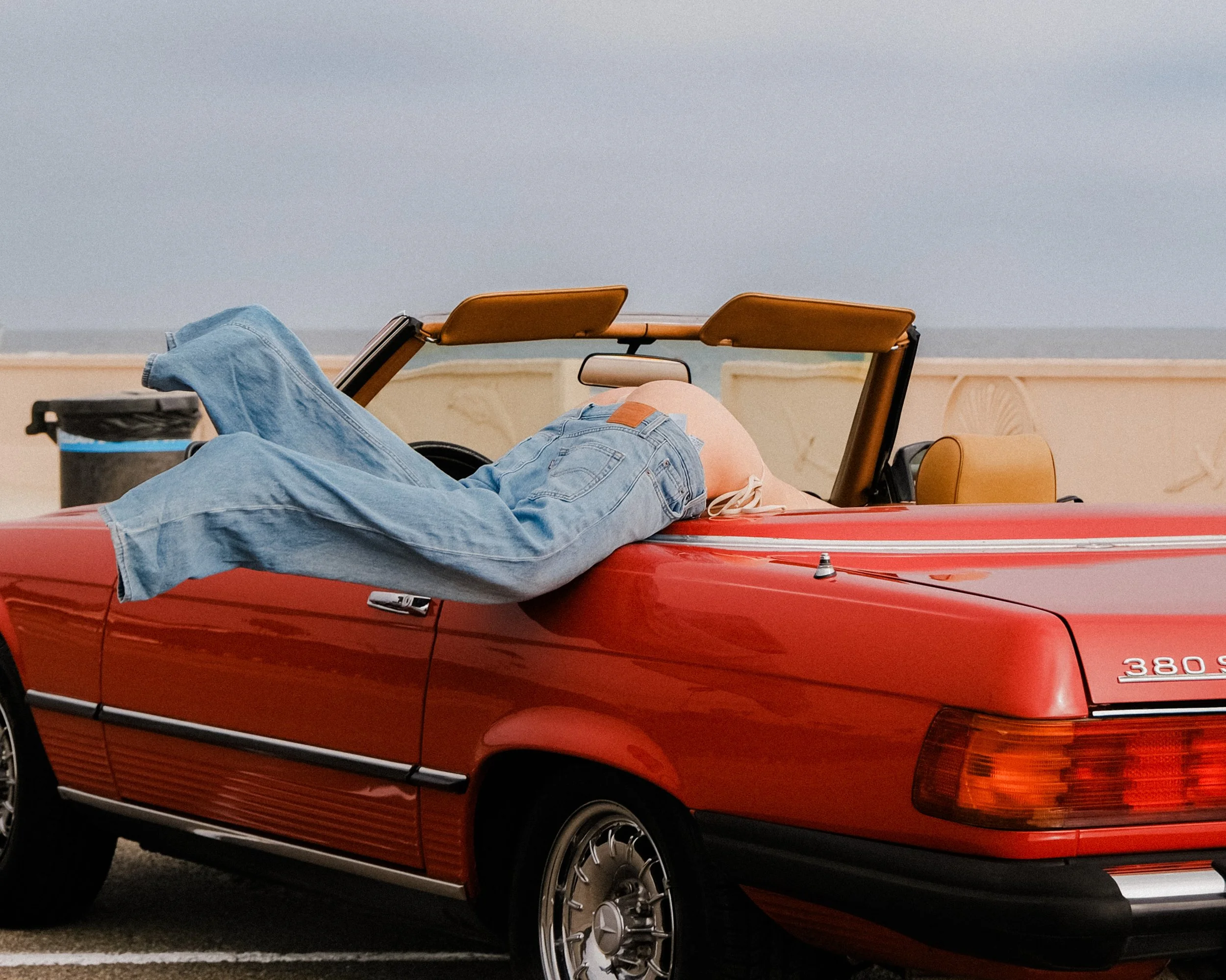 A person lying on a red convertible car with the top down, wearing blue jeans and a light-colored top, with ocean and cloudy sky in the background.