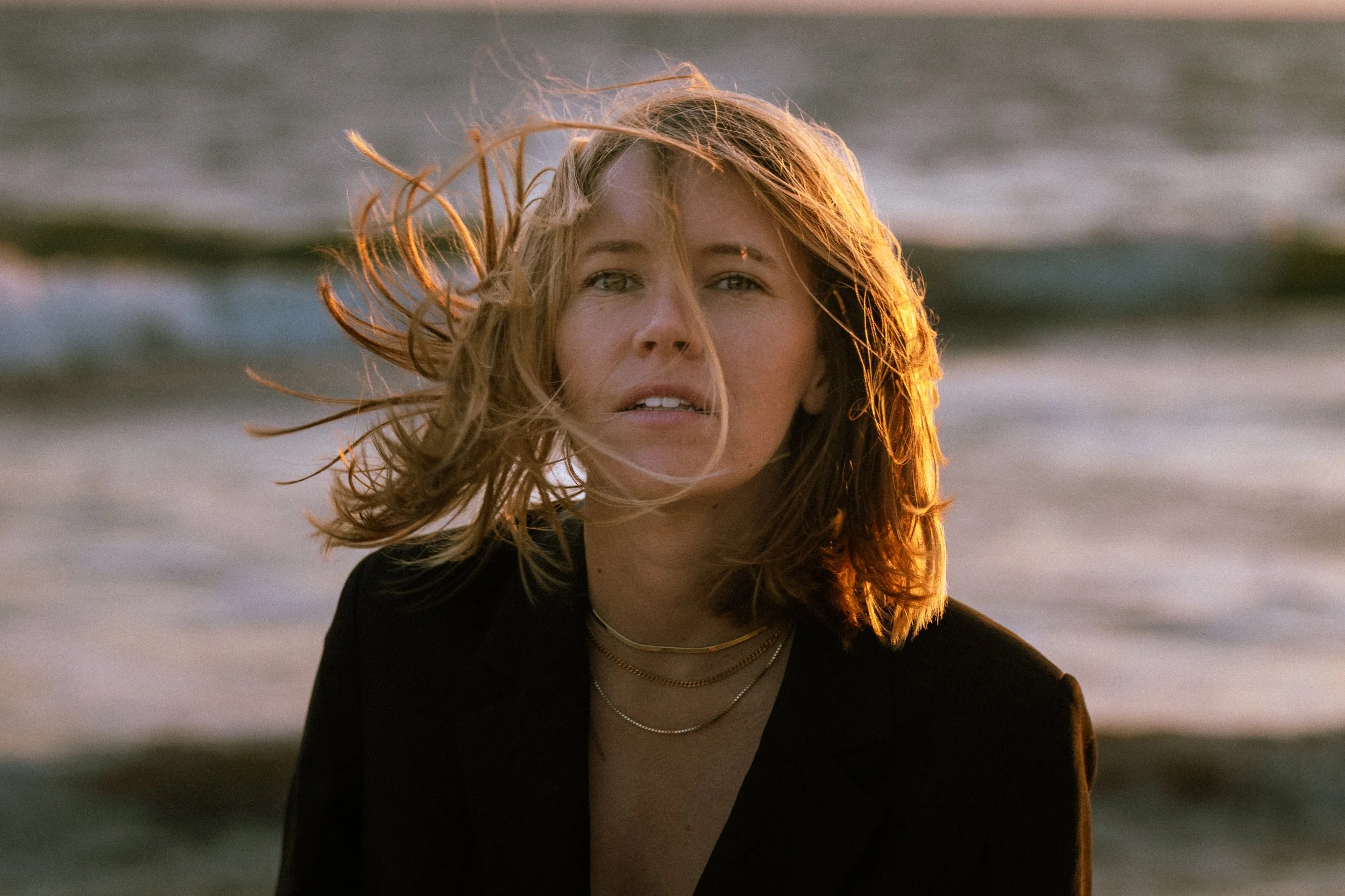 Young woman with wavy blonde hair standing on the beach with the ocean in the background, illuminated by sunset light.