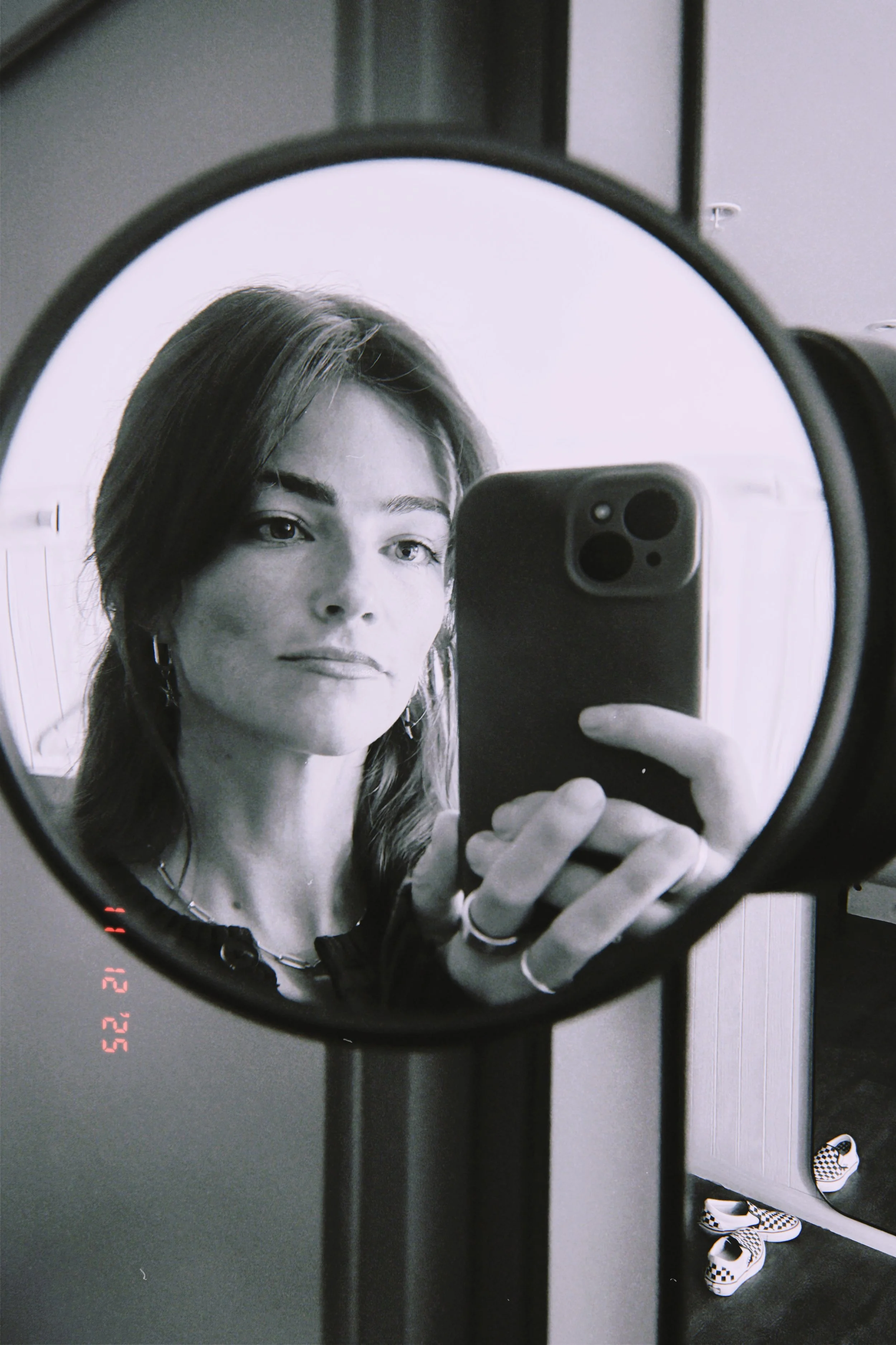 A woman taking a mirror selfie with a smartphone, showing her face and shoulder. She has light-colored hair, earrings, and a necklace. The reflection is in an oval mirror, and there are checkered Vans shoes on the floor.