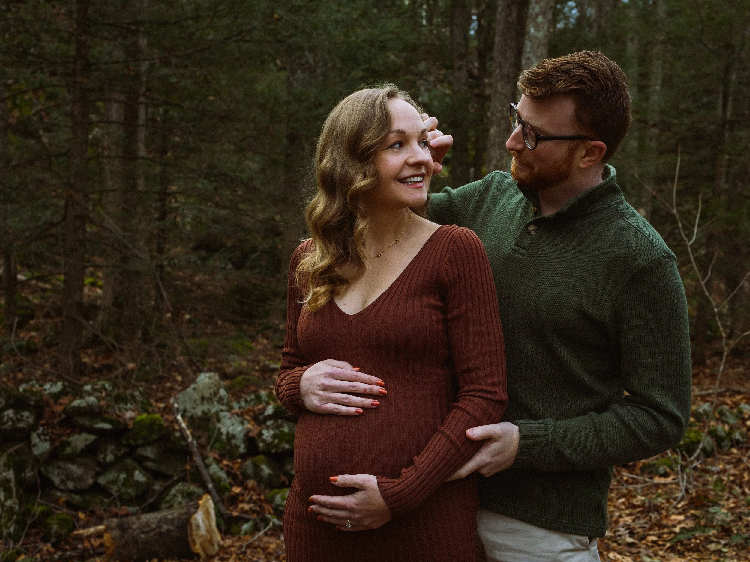 A couple standing close together in a wooded area, with the man's arm around the woman's shoulder and their hands touching her pregnant belly, smiling and looking at each other.