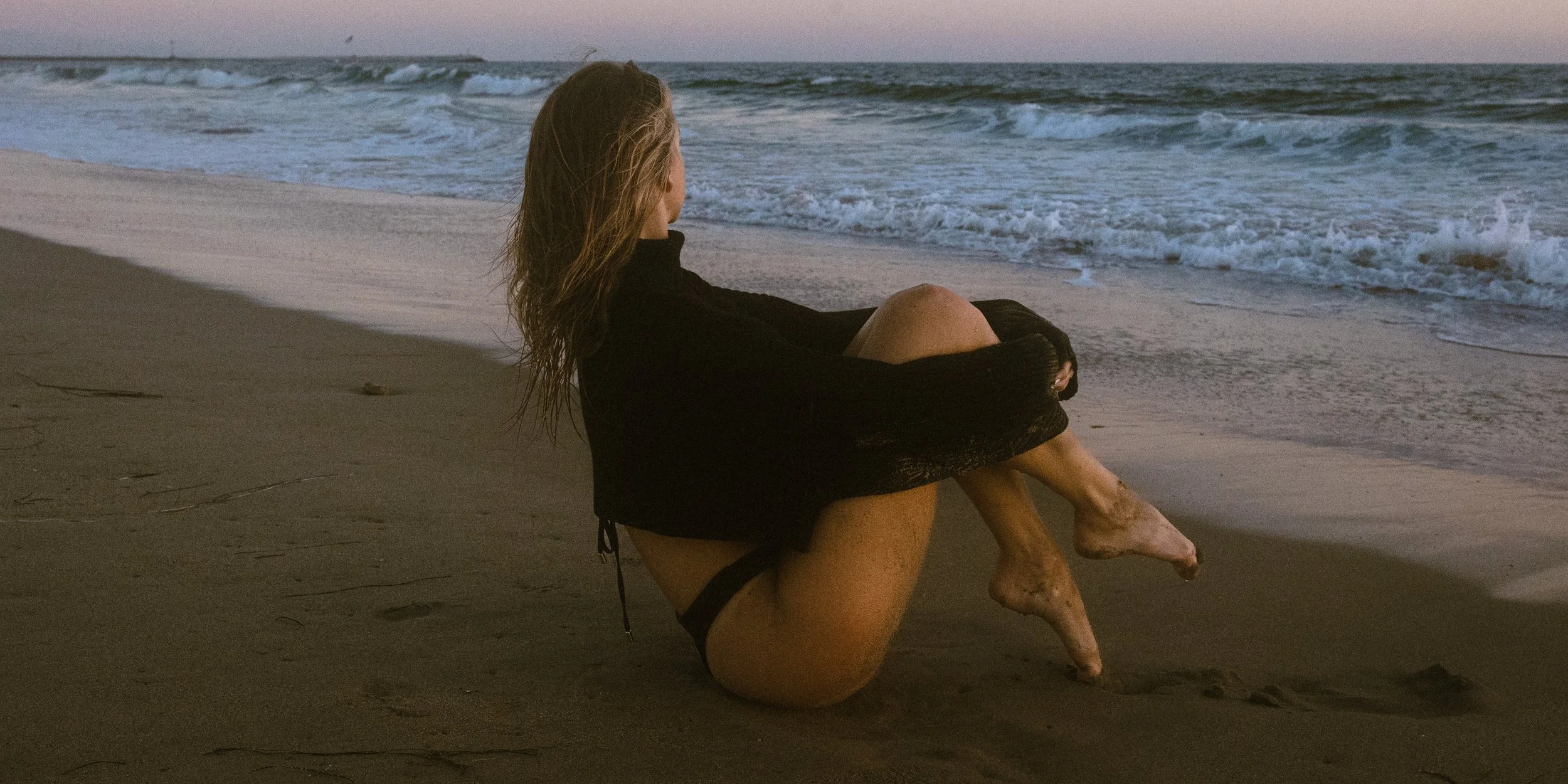 A woman sitting on the beach at sunset, looking out at the ocean waves.