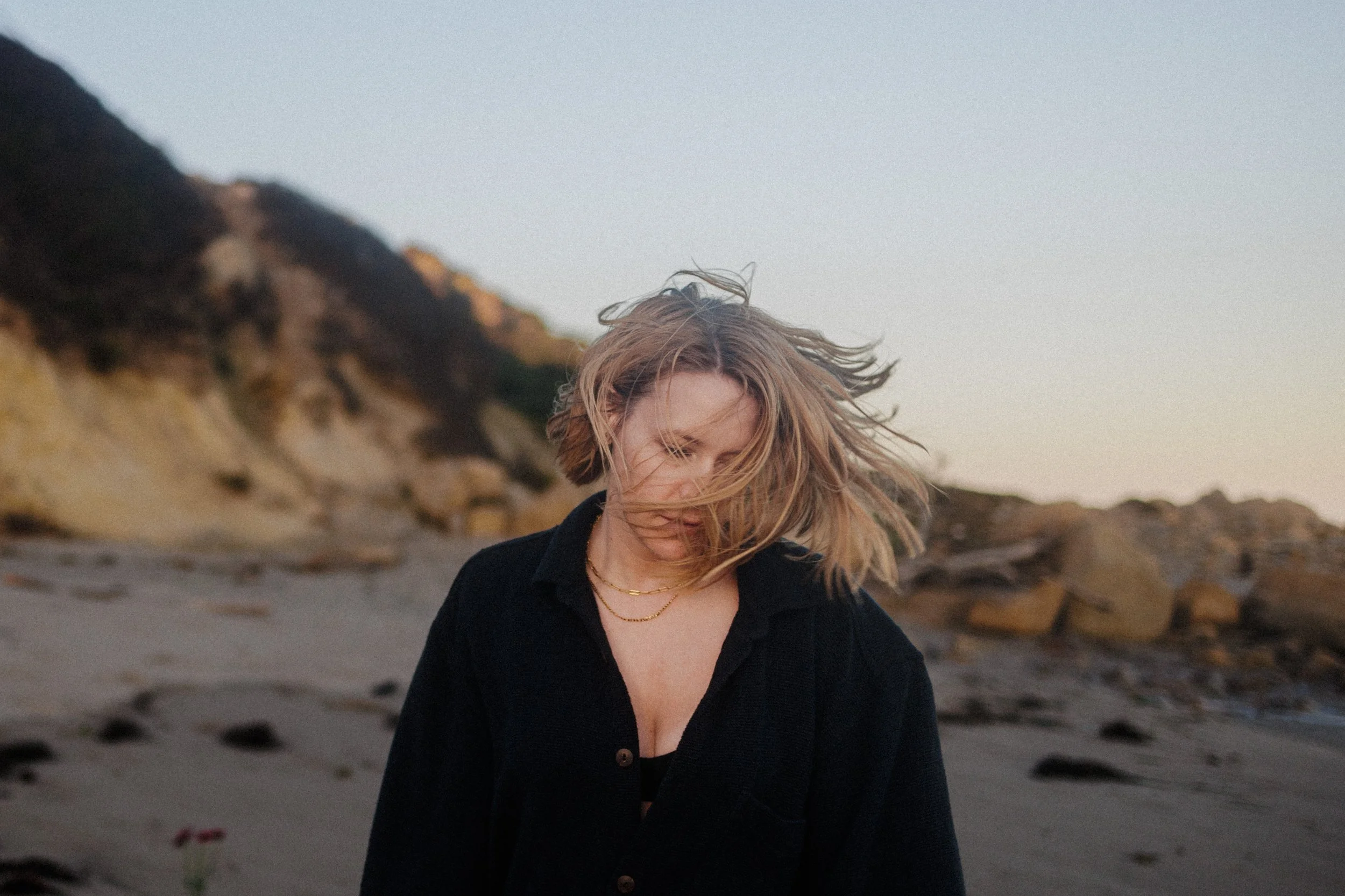 A young woman with blonde hair standing outdoors on a beach with rocks, wearing a black jacket and layered necklaces, looking down with wind blowing her hair, during sunset or dusk.