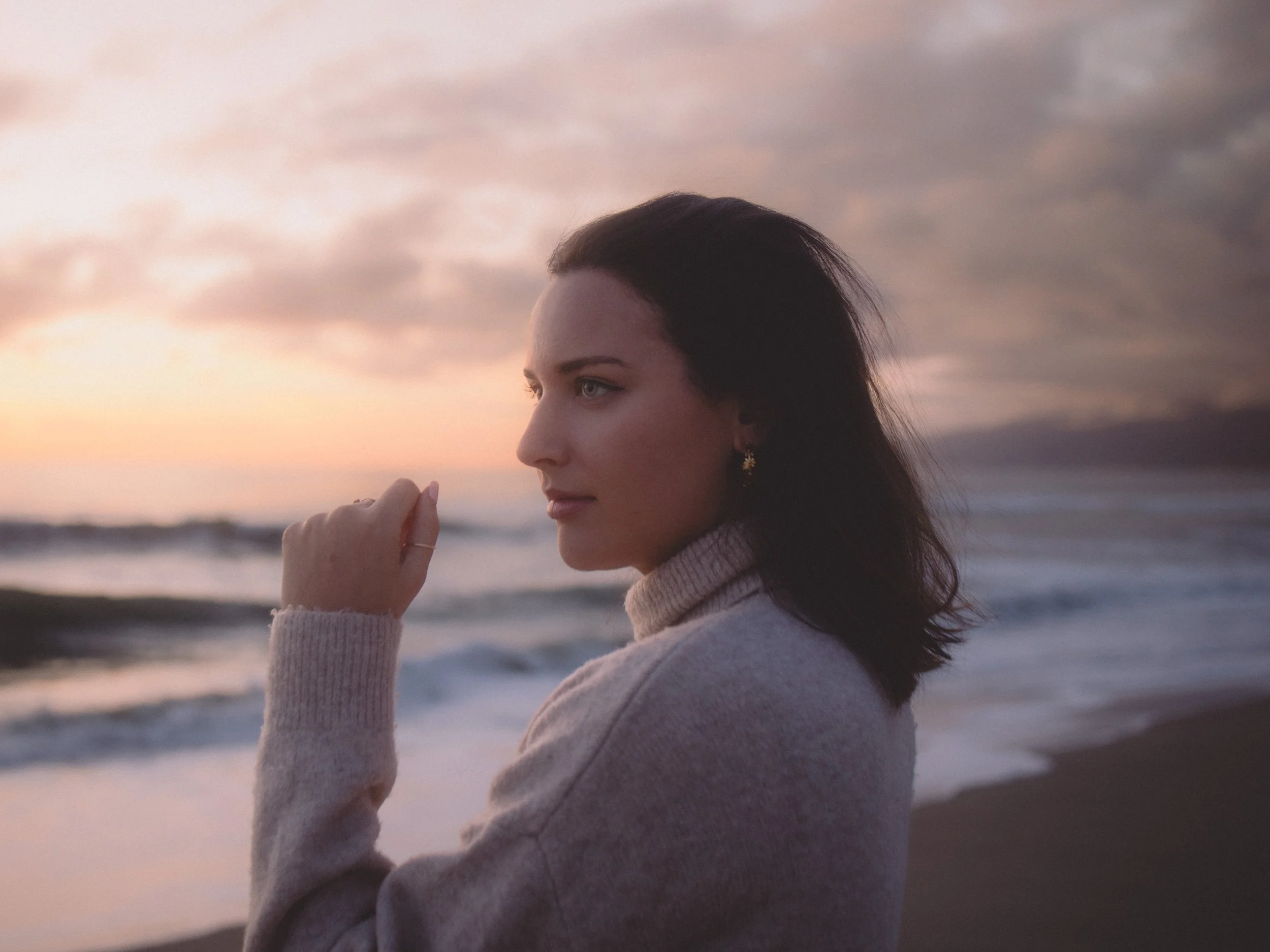 A woman with dark hair and light blue eyes looking away, with a sunset sky and ocean in the background.