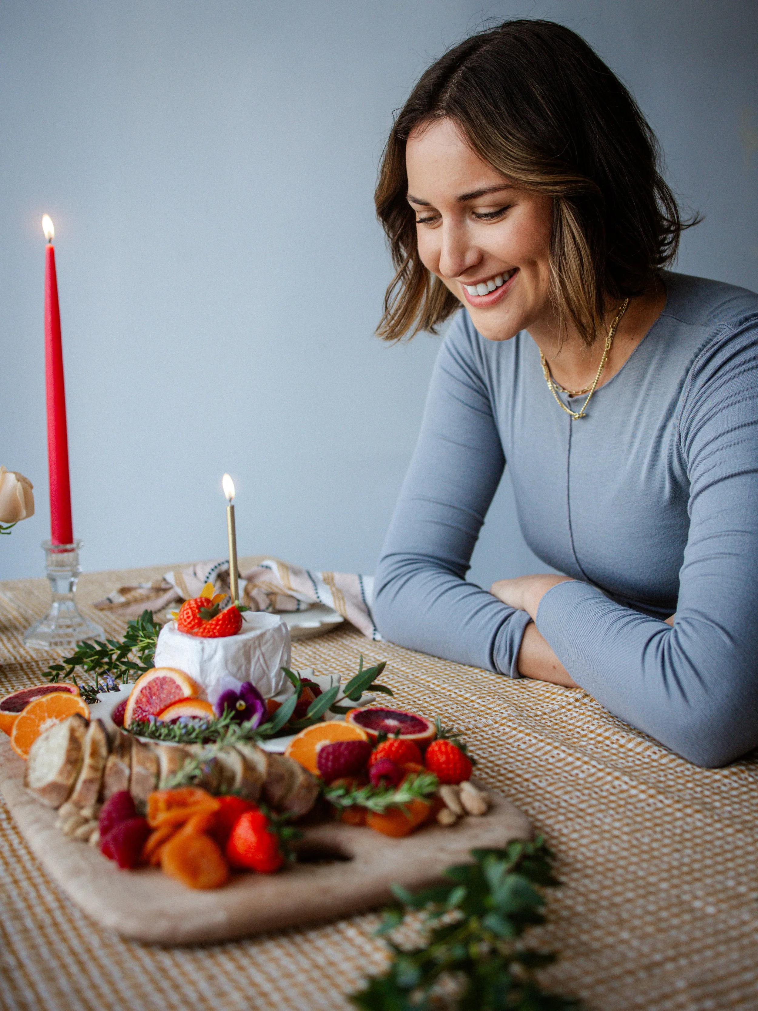 A woman with short dark hair smiling at a table with a variety of colorful food, including a small cake with strawberries and whipped cream, and a spread of fruits and other dishes, during a celebration.