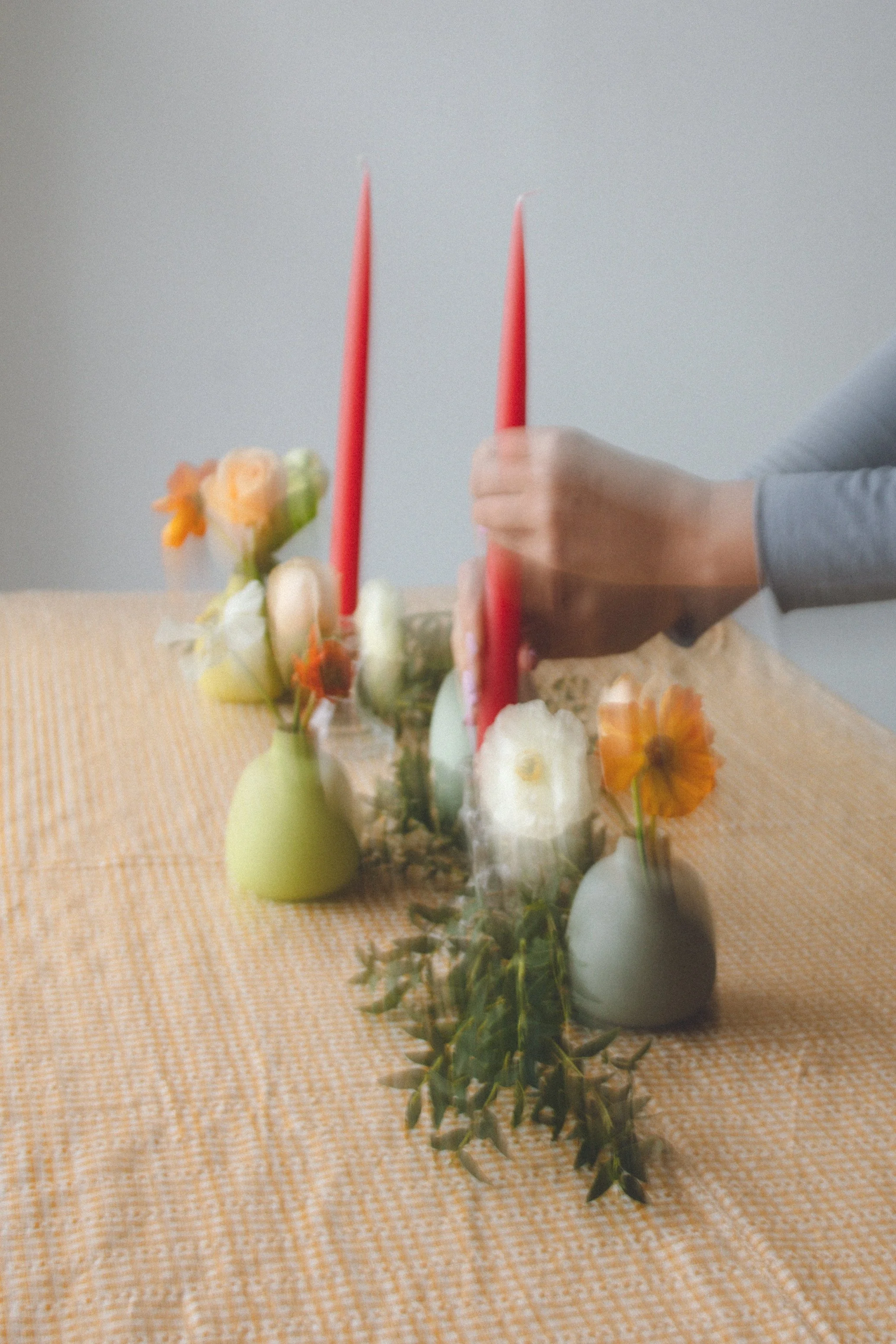 A table with a floral arrangement, two tall red candles, and decorative objects in plain vases, all on a light orange and white checkered tablecloth.