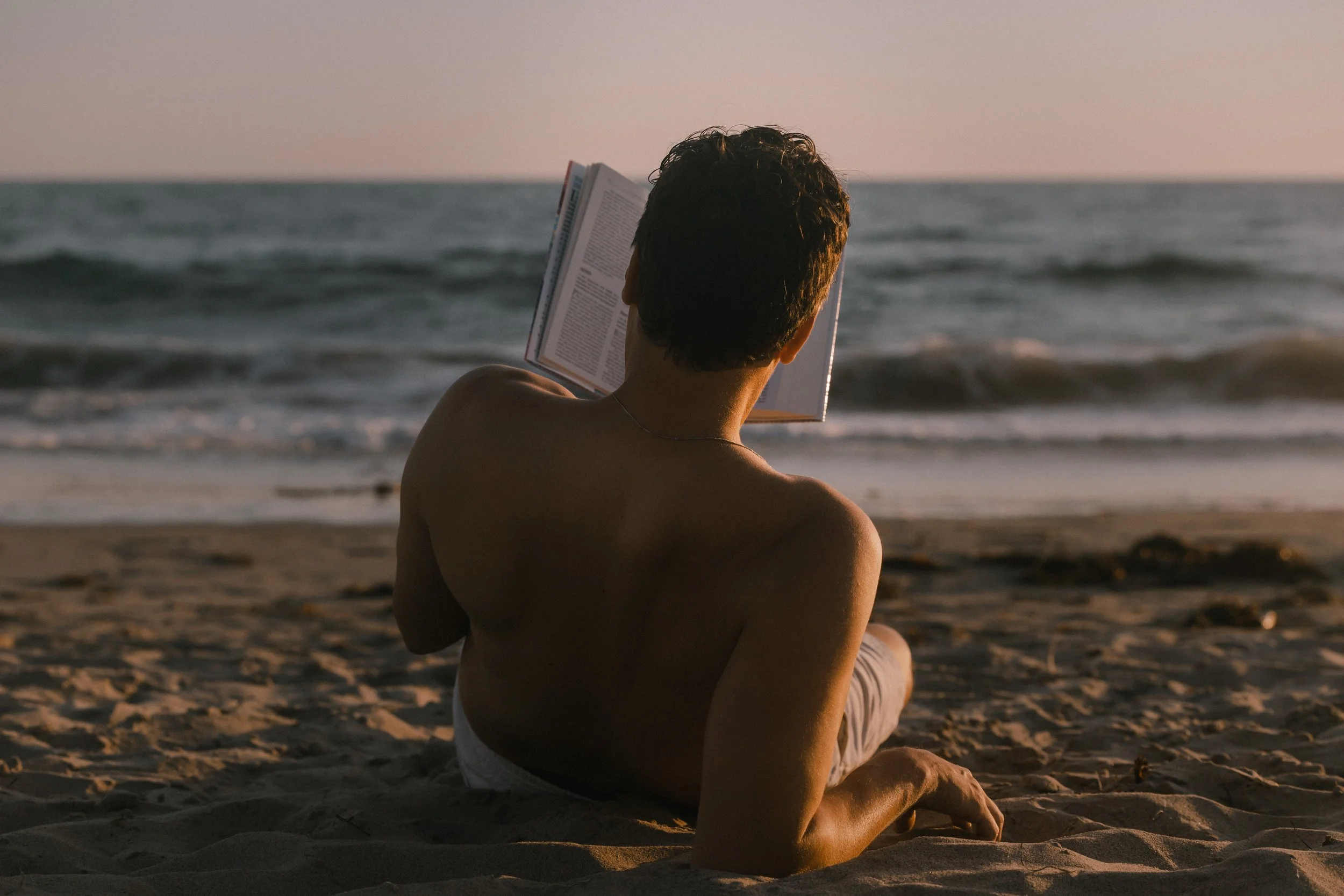 Person sitting on the beach reading a book during sunset.