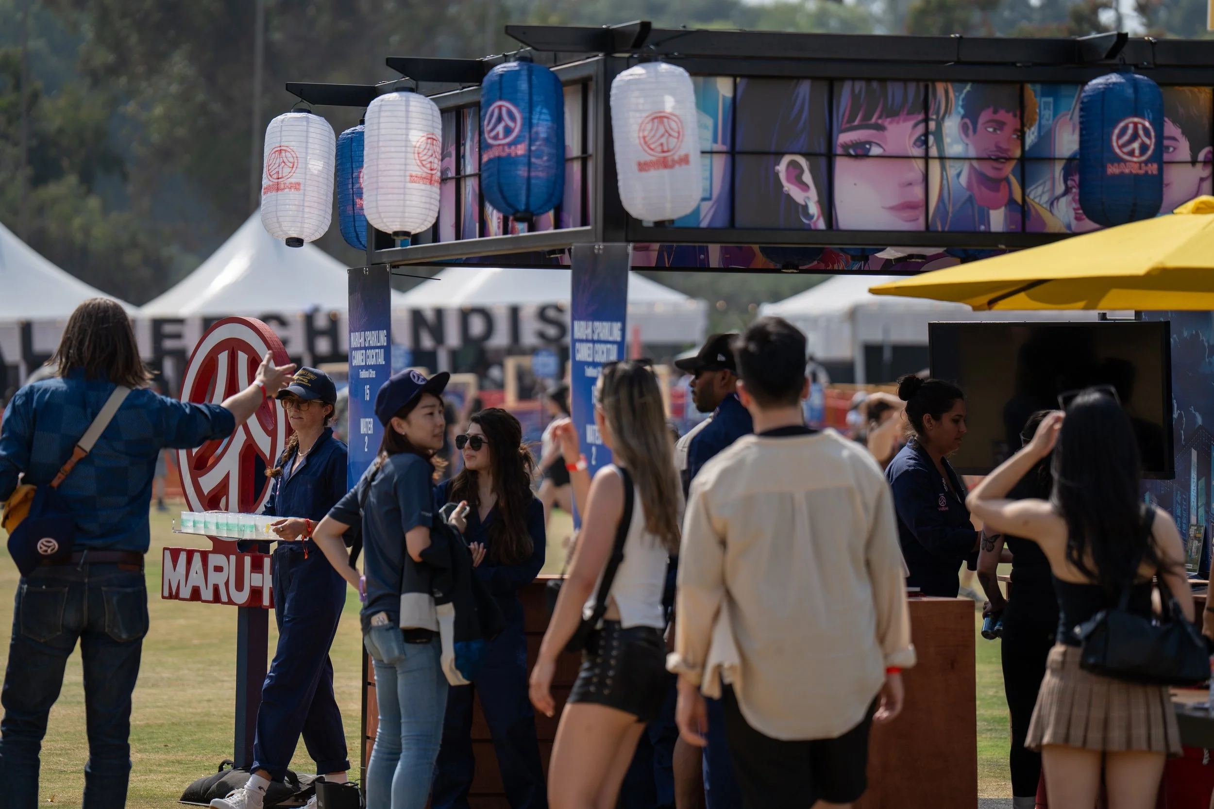 People gather at an outdoor event with booths, banners, and lanterns, and a large colorful mural of animated characters in the background.